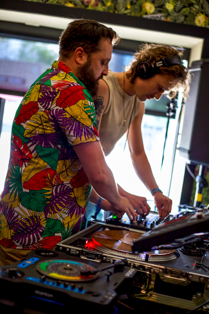 Two people DJing with turntables and equipment indoors, one wearing a colorful floral shirt, the other with curly hair.