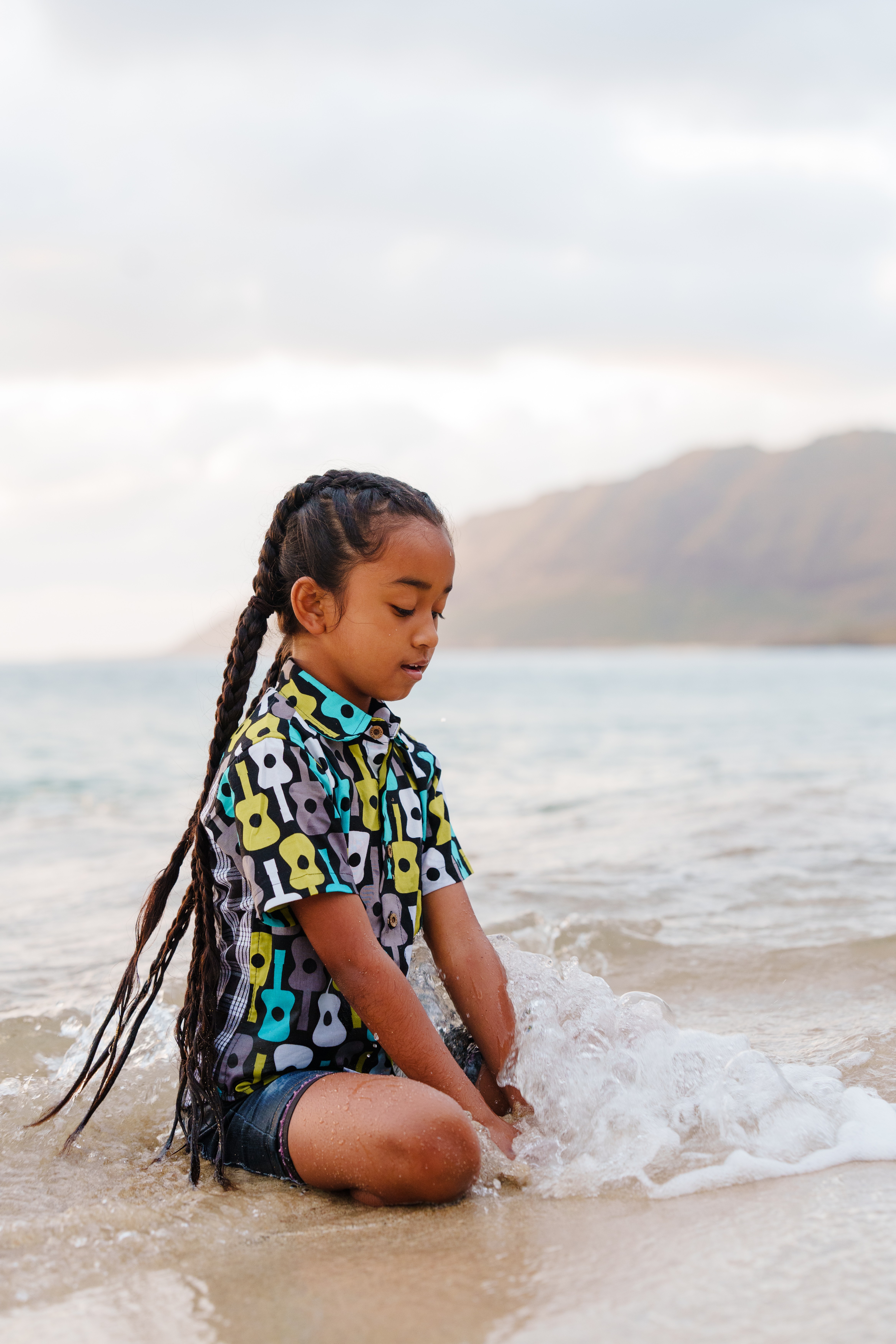 Child with long braided hair sitting in shallow water at the beach, wearing a colorful patterned shirt, with mountains and cloudy sky in background.