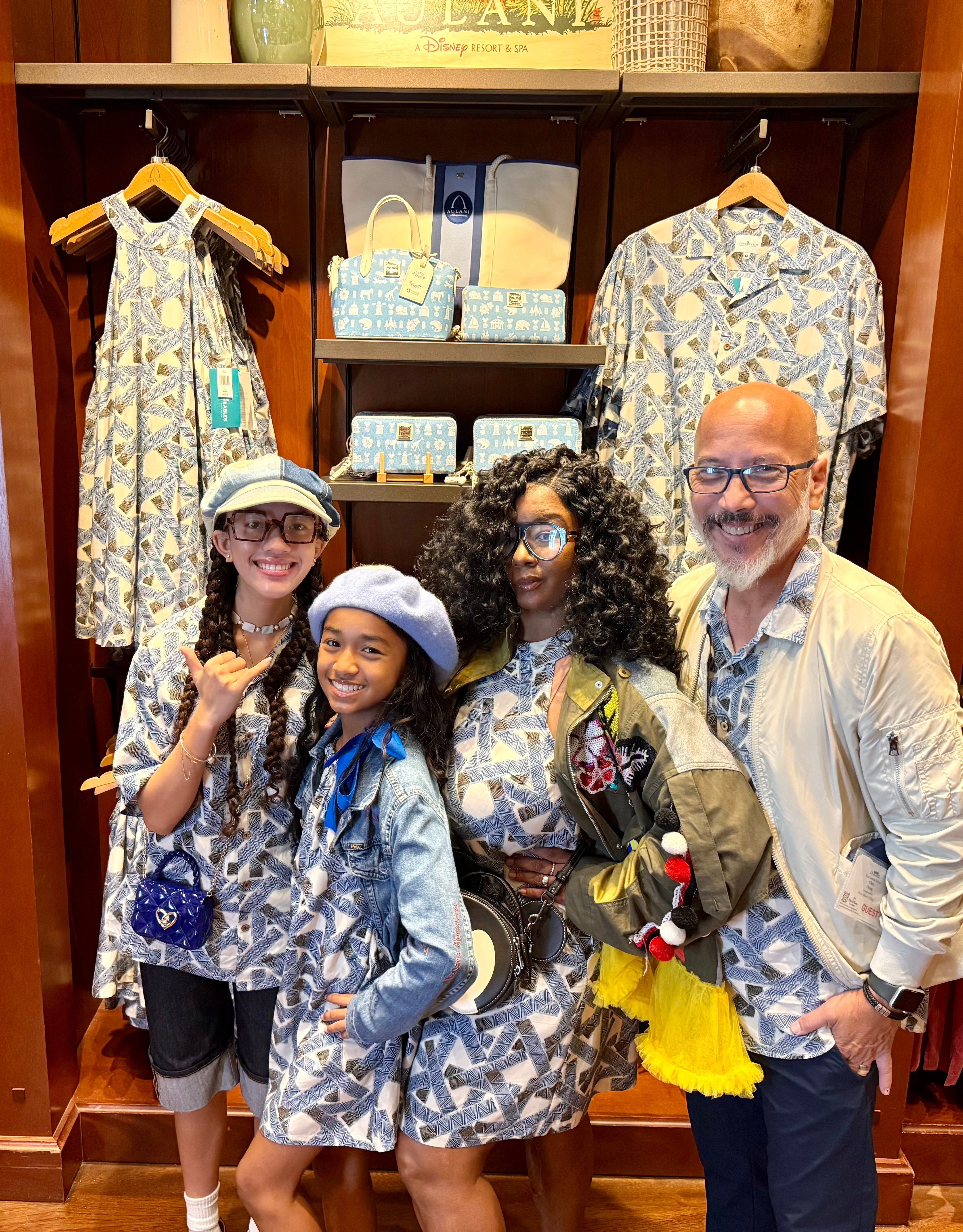 Four people, two children and two adults, stand in front of a clothing display with patterned dresses hanging behind them. They are smiling and posing for the photo.