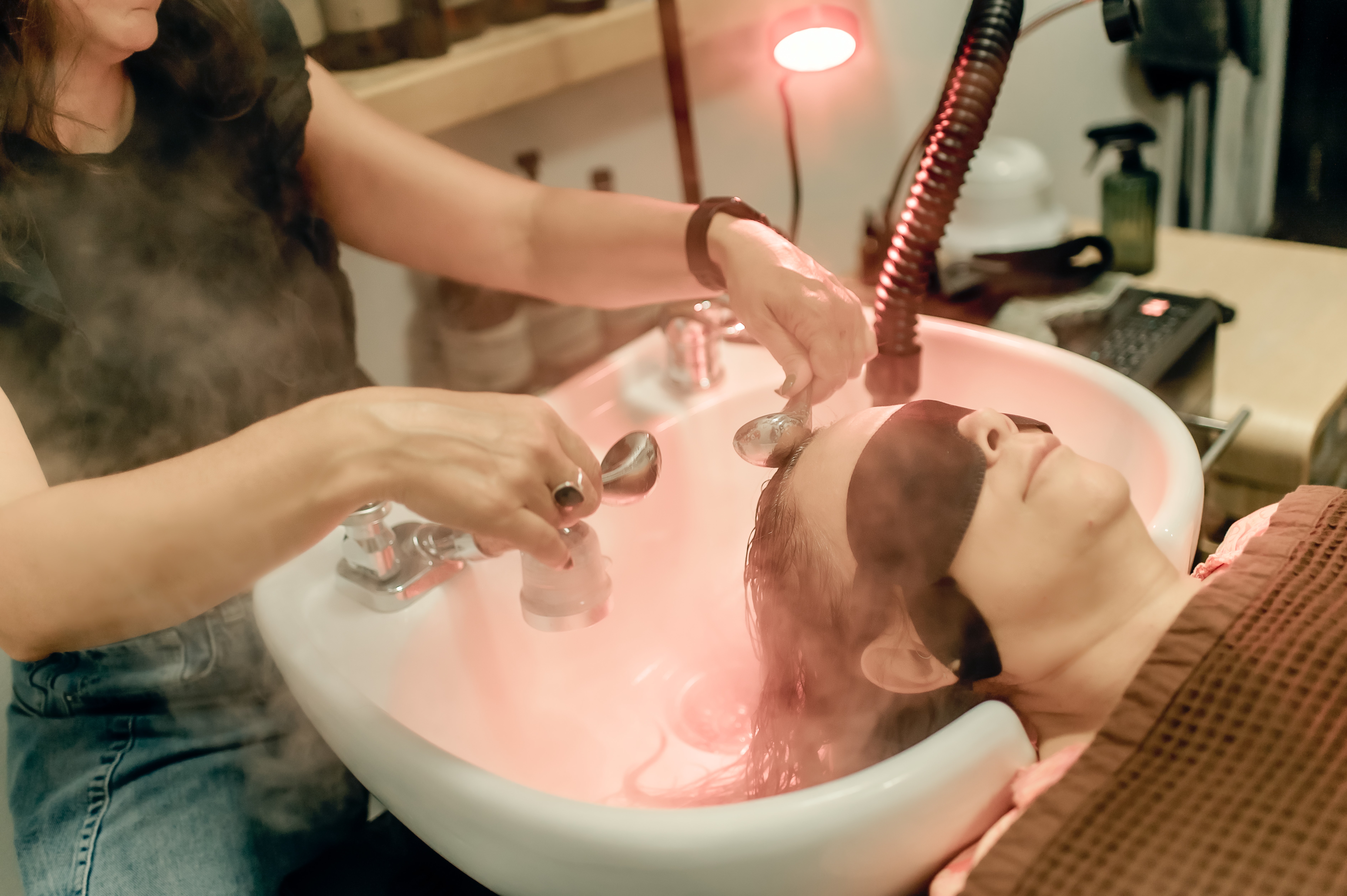 Person receiving a facial treatment with steam and a professional applying a mask or cleanser, in a salon setting.