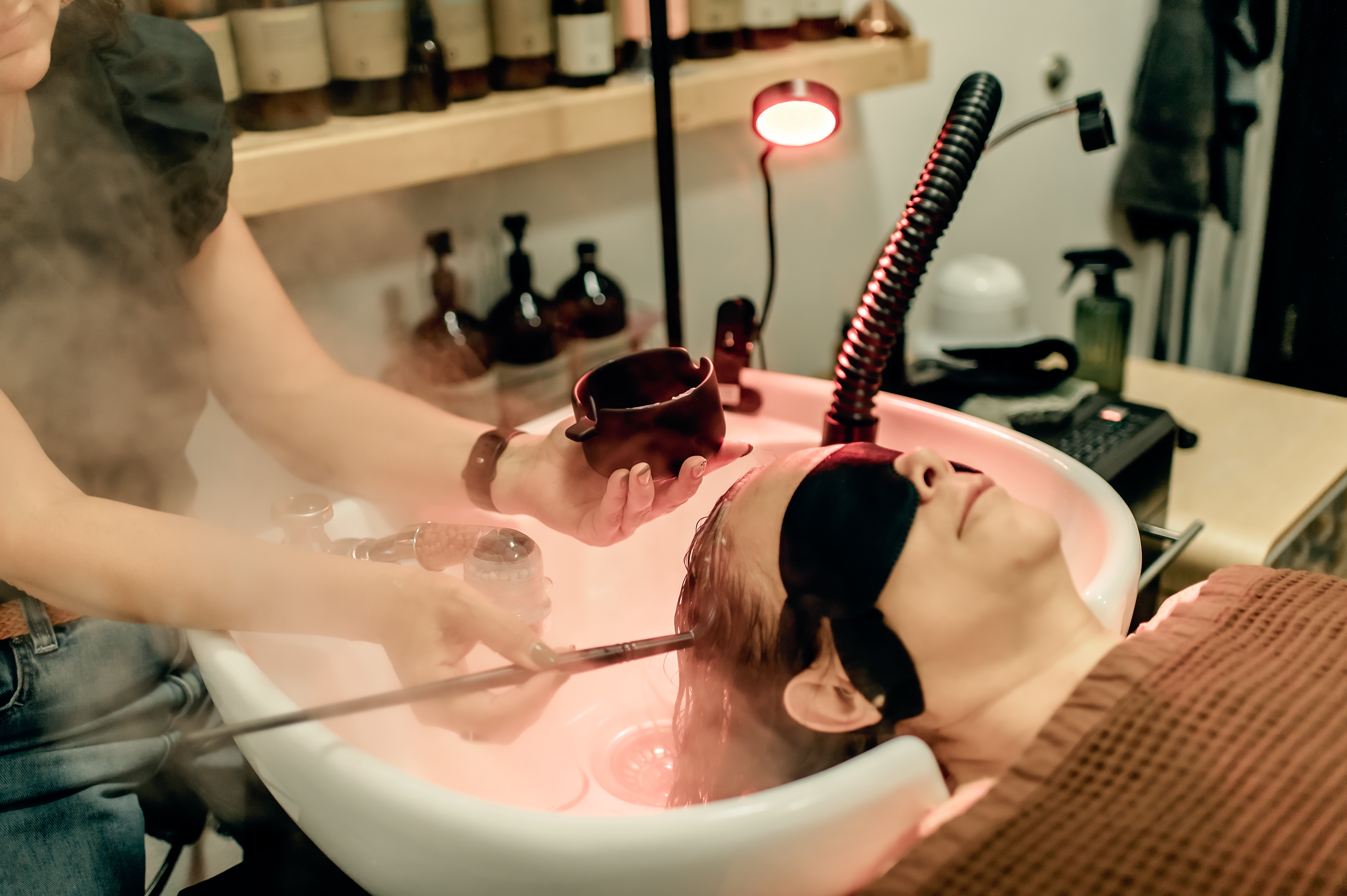 Person with head in a sink, receiving a hair treatment, with another person holding a bowl and tool nearby, in a salon setting.
