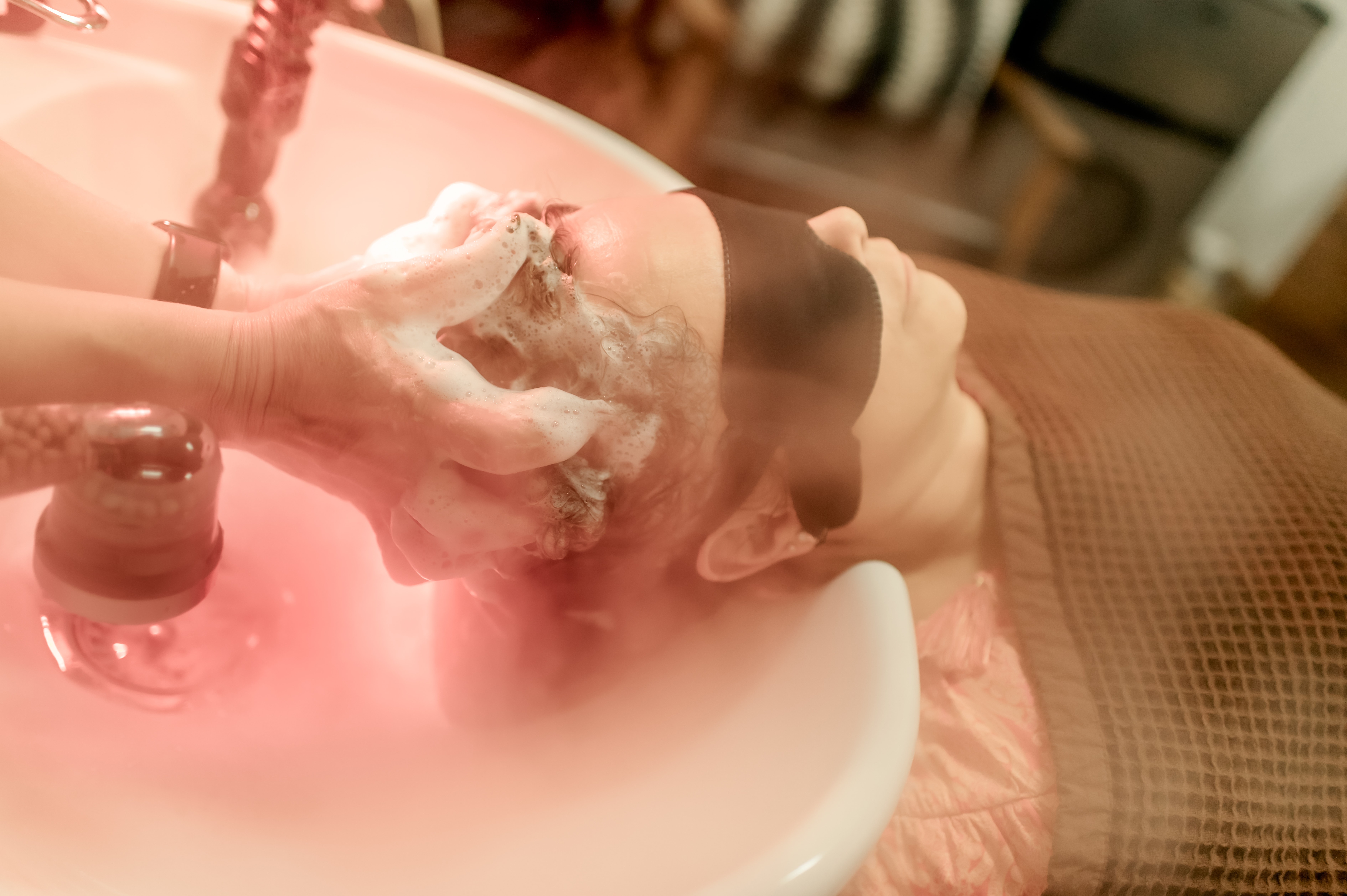 Person lying in a bathtub with soap bubbles, with a hand reaching towards their face, pink lighting, and a wooden surface nearby.