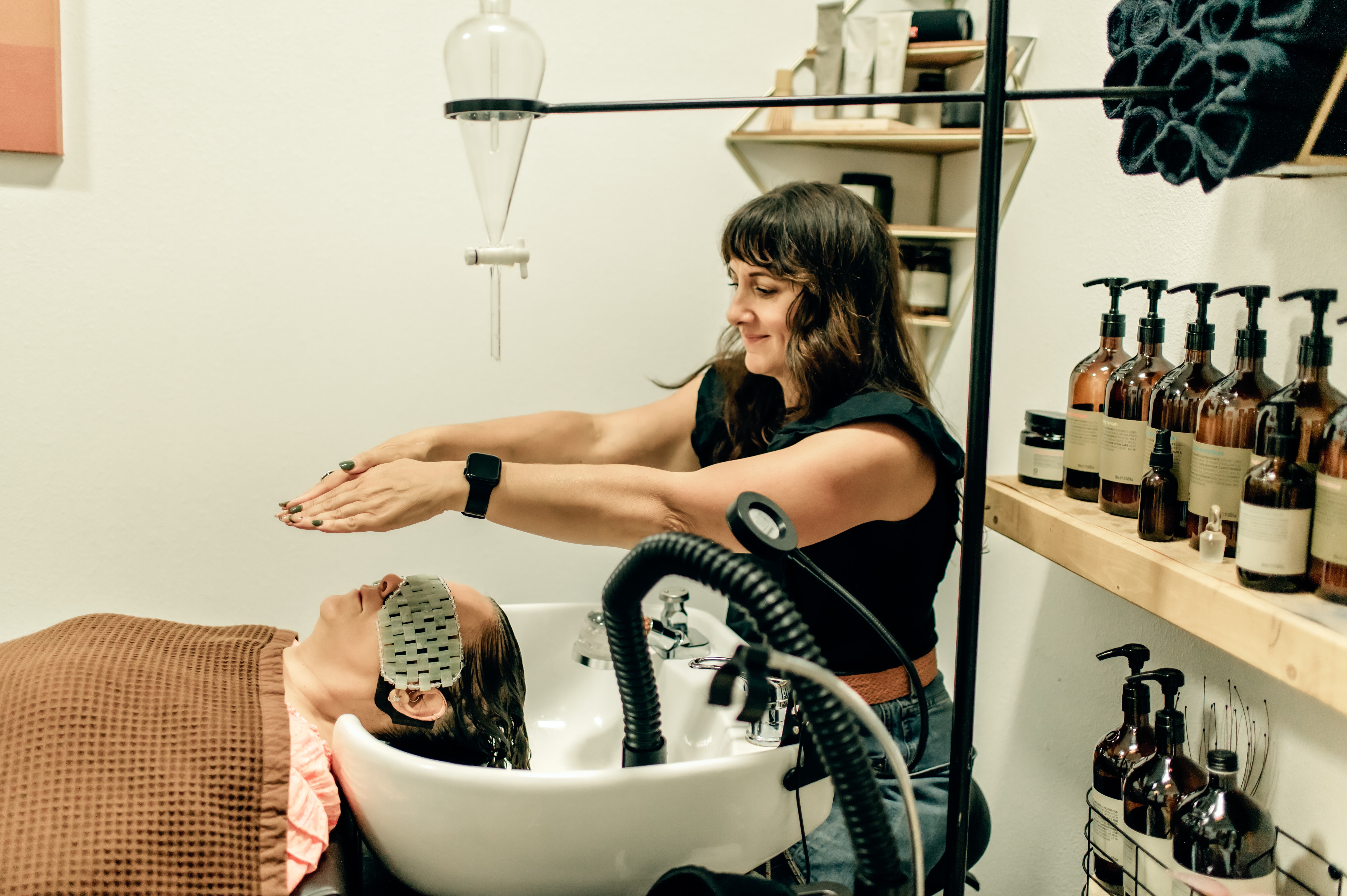 Woman with dark hair stretching her arms in a salon or spa, with bottles on shelves behind her.