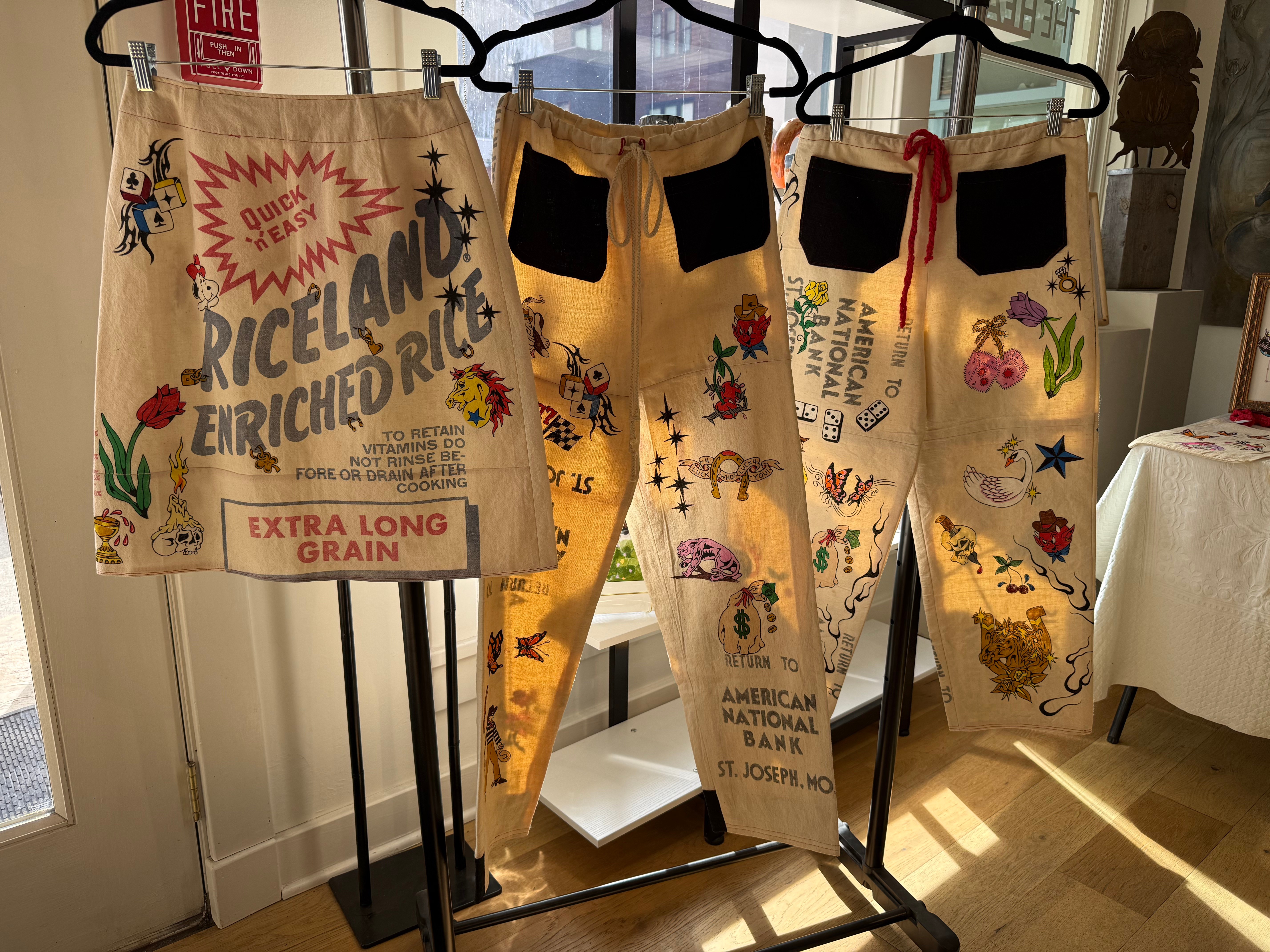 Three pairs of pants with colorful embroidered designs hang on a clothing rack, sunlight streaming in from a window.