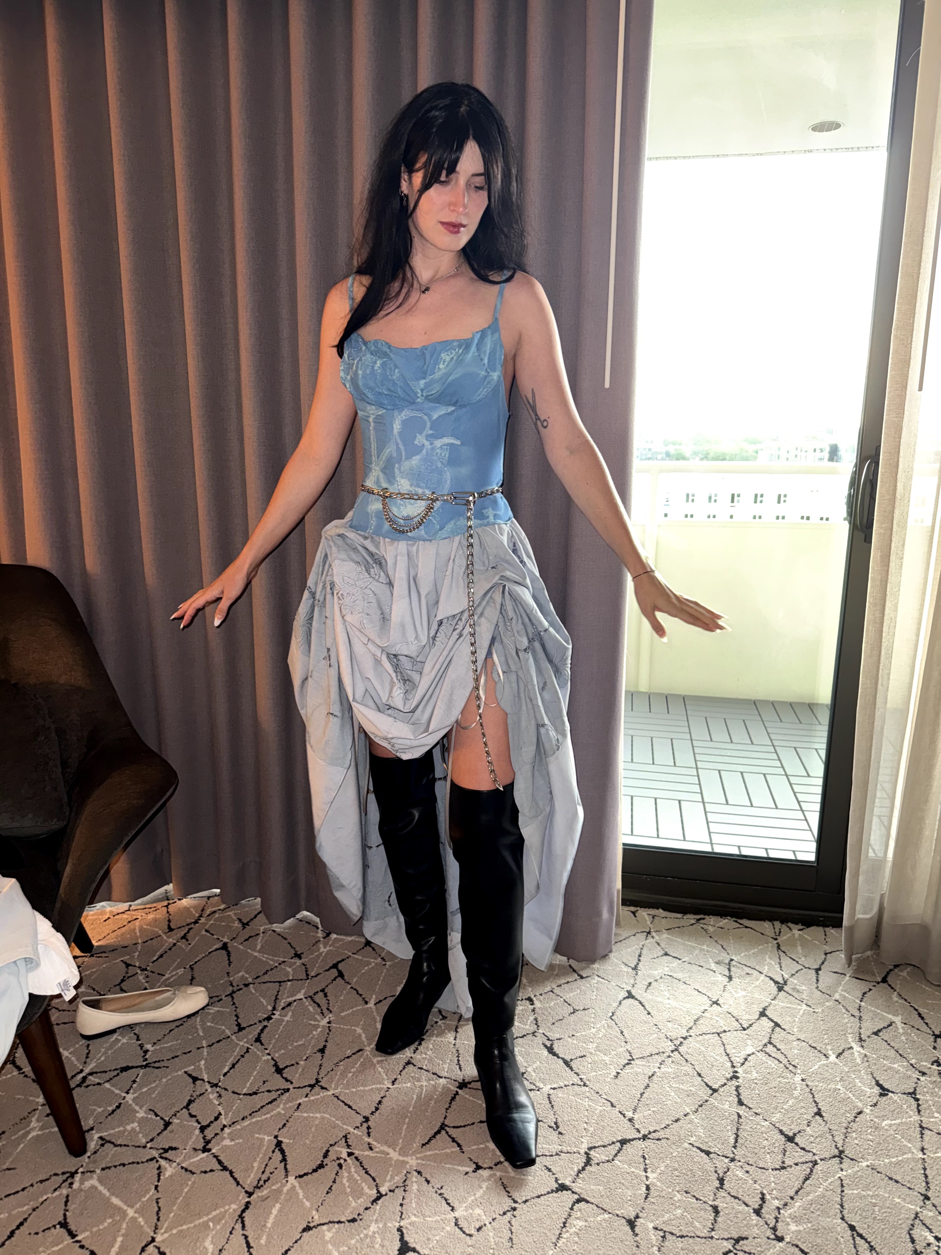 Woman in a blue top and high boots standing indoors near a window and curtain, with a patterned carpet and furniture nearby.