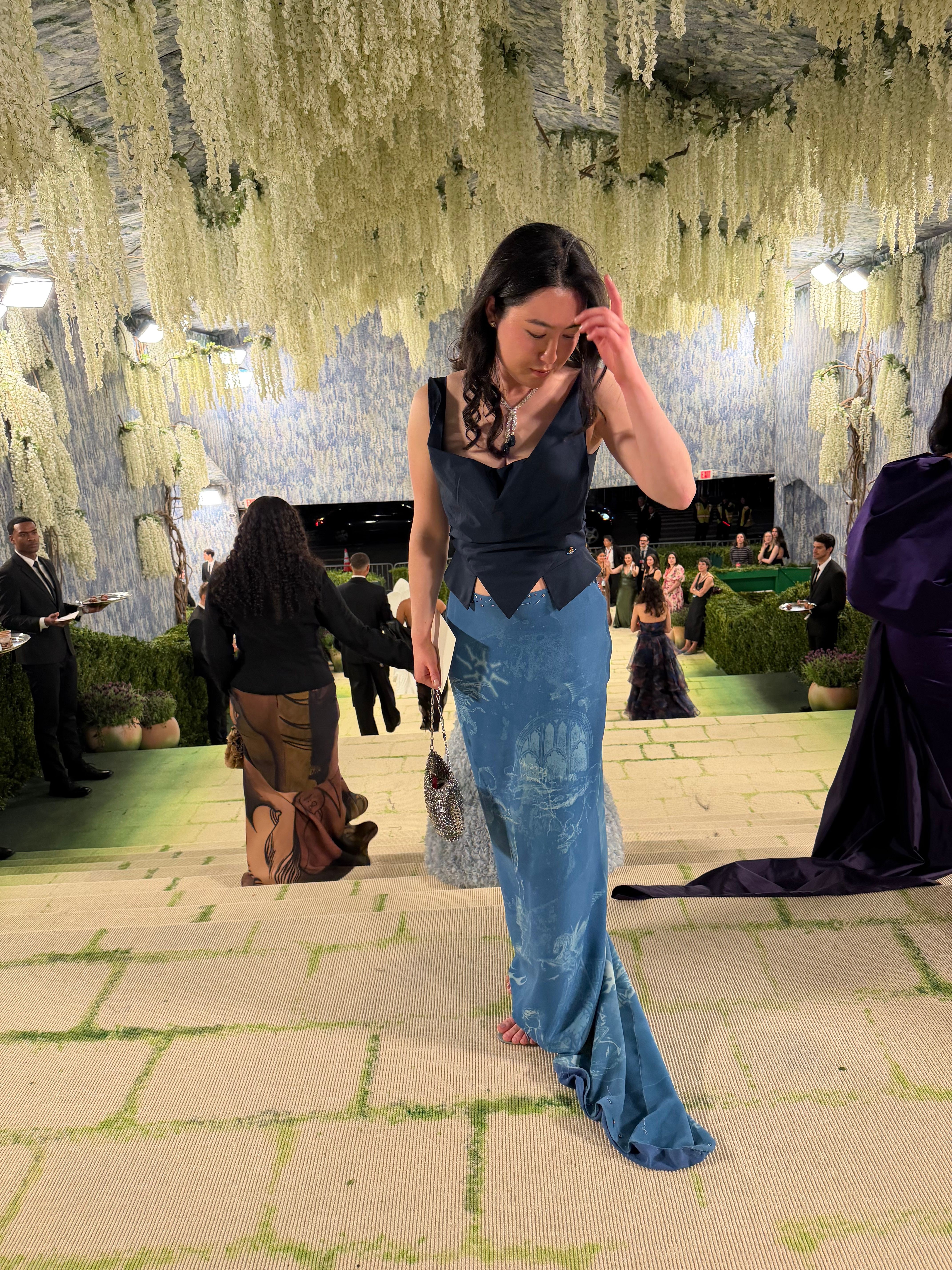 Woman with long dark hair in a black top and blue jeans walking outdoors near a water fountain, trees, and other people.