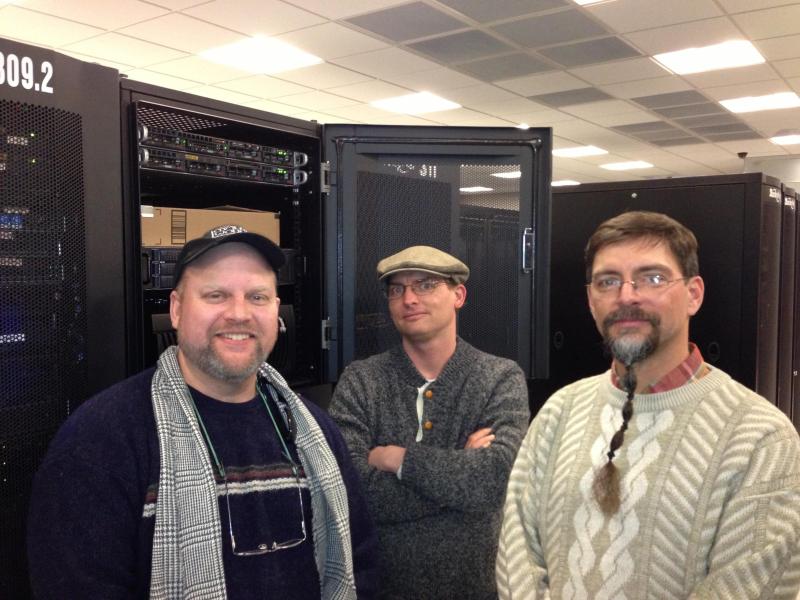 Three men standing in front of server racks in a room with a ceiling grid and fluorescent lights.