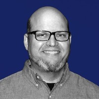 A smiling man with glasses and a beard, wearing a button-up shirt, against a blue background.