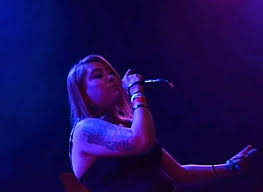 Person singing into a microphone on stage with colorful lighting, holding the microphone with one hand and gesturing with the other.