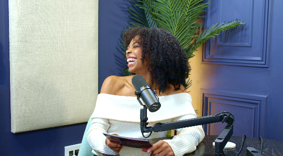Woman with curly hair smiling, holding a notebook, sitting in a recording studio with a microphone, green plant, and blue wall.