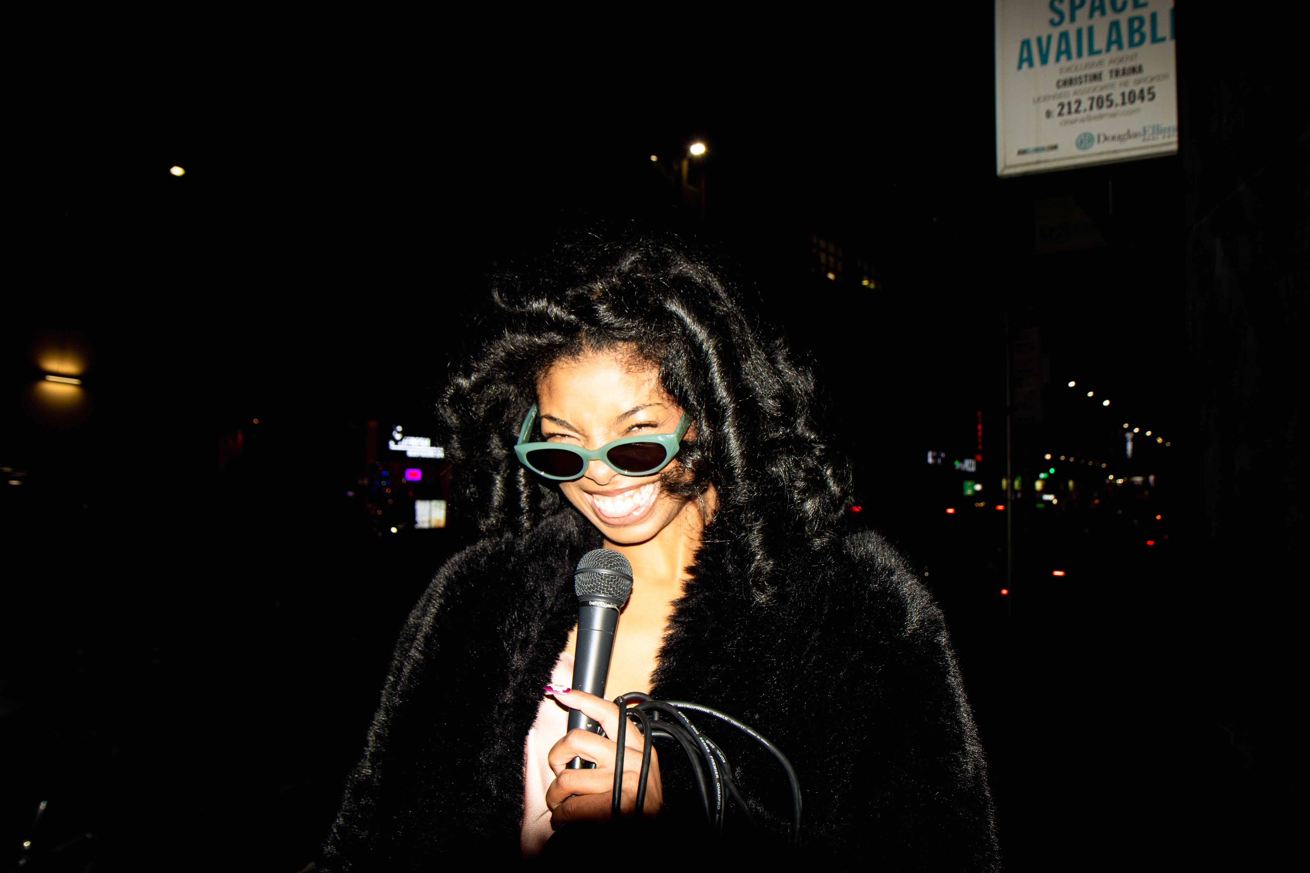 Person with curly hair wearing sunglasses and a black coat, holding a microphone outdoors at night.