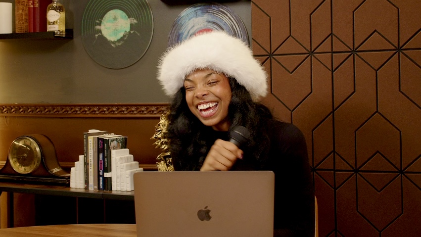 Woman wearing a white fluffy hat smiling at a laptop in a warmly decorated room.
