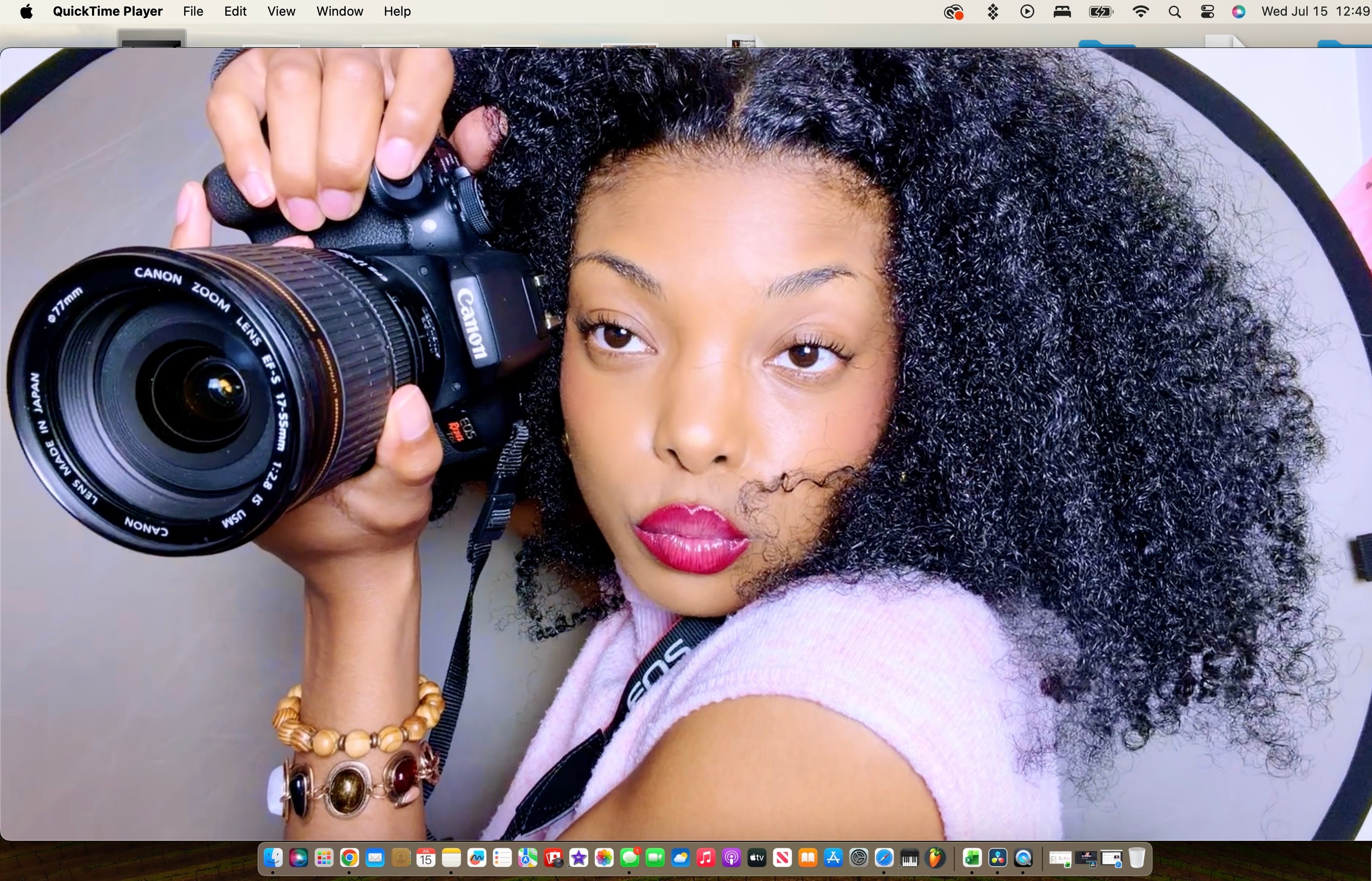 Young woman with curly hair holding a camera, looking at the viewer, wearing a pink top and jewelry.