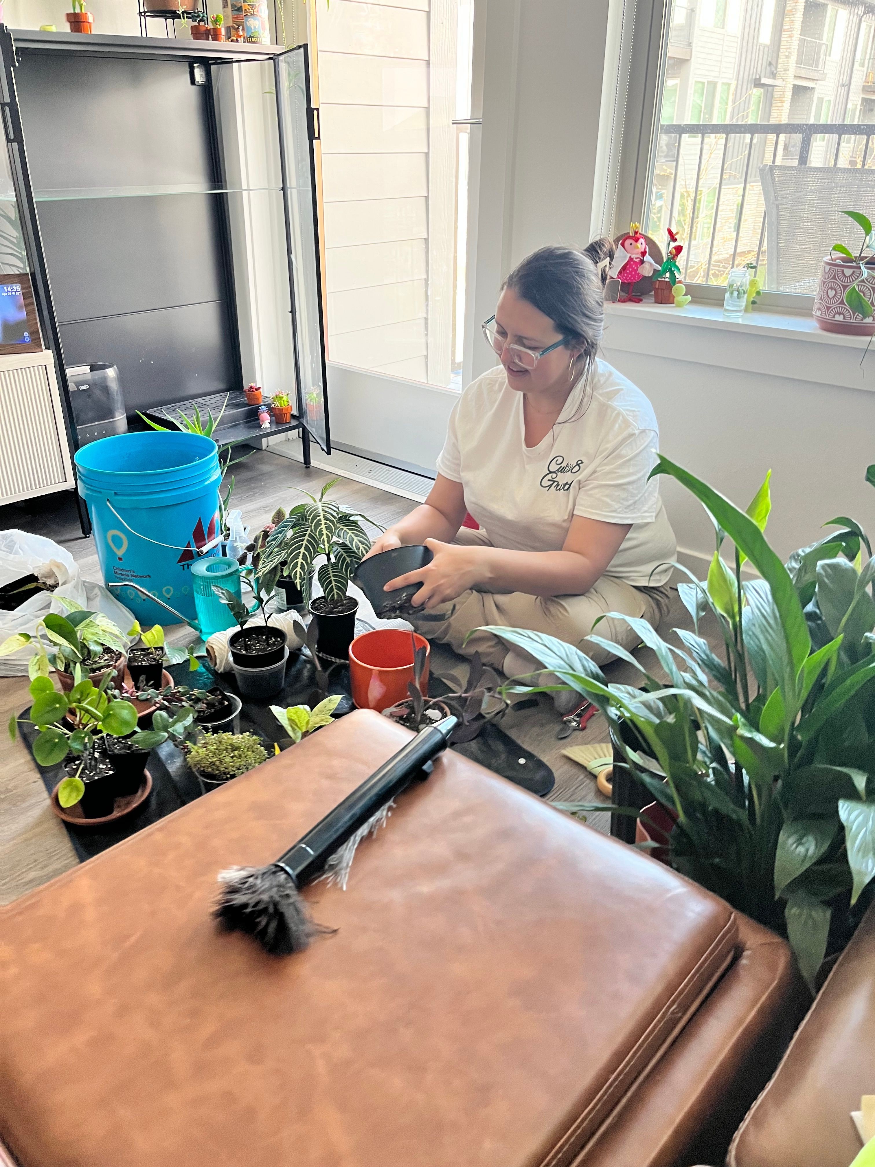 Woman with glasses and white shirt tending to plants on a windowsill, surrounded by gardening tools and colorful pots.