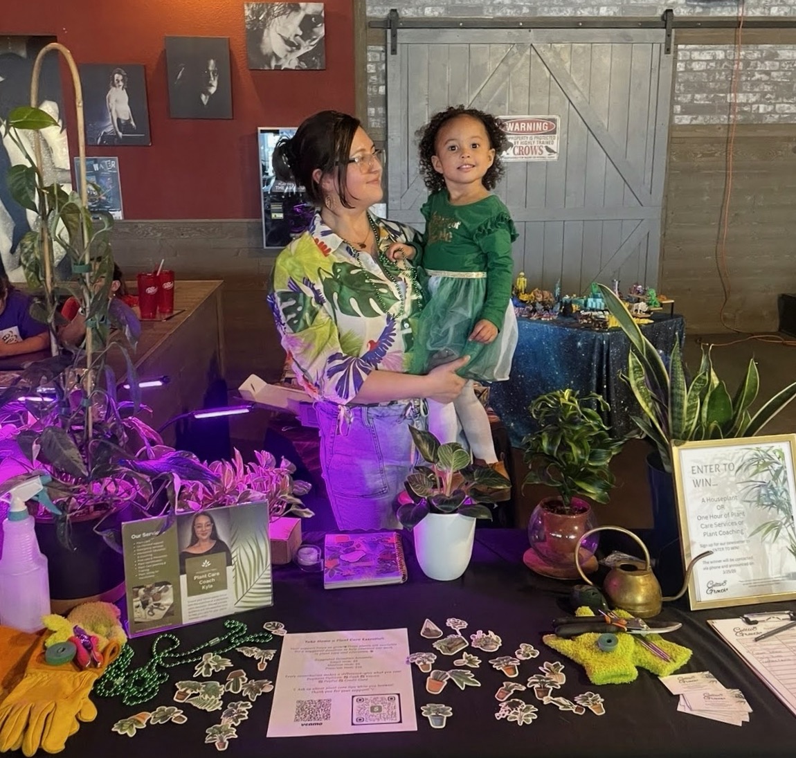 Woman holding a young girl at a table with plants, photos, and informational materials, in a decorated indoor space.