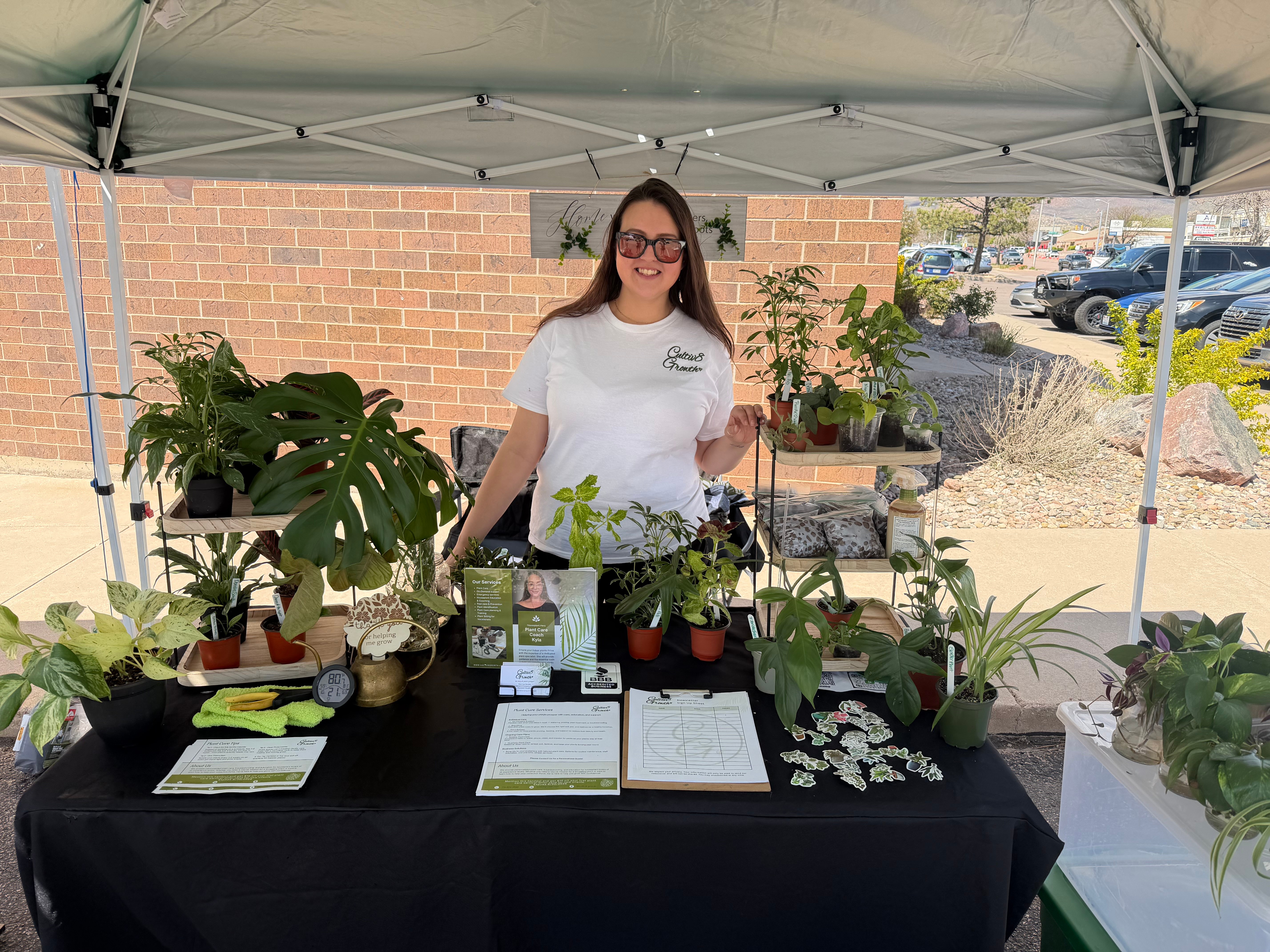 Woman standing behind a table with potted plants under a canopy, brick wall in background, smiling, wearing glasses and a white shirt.
