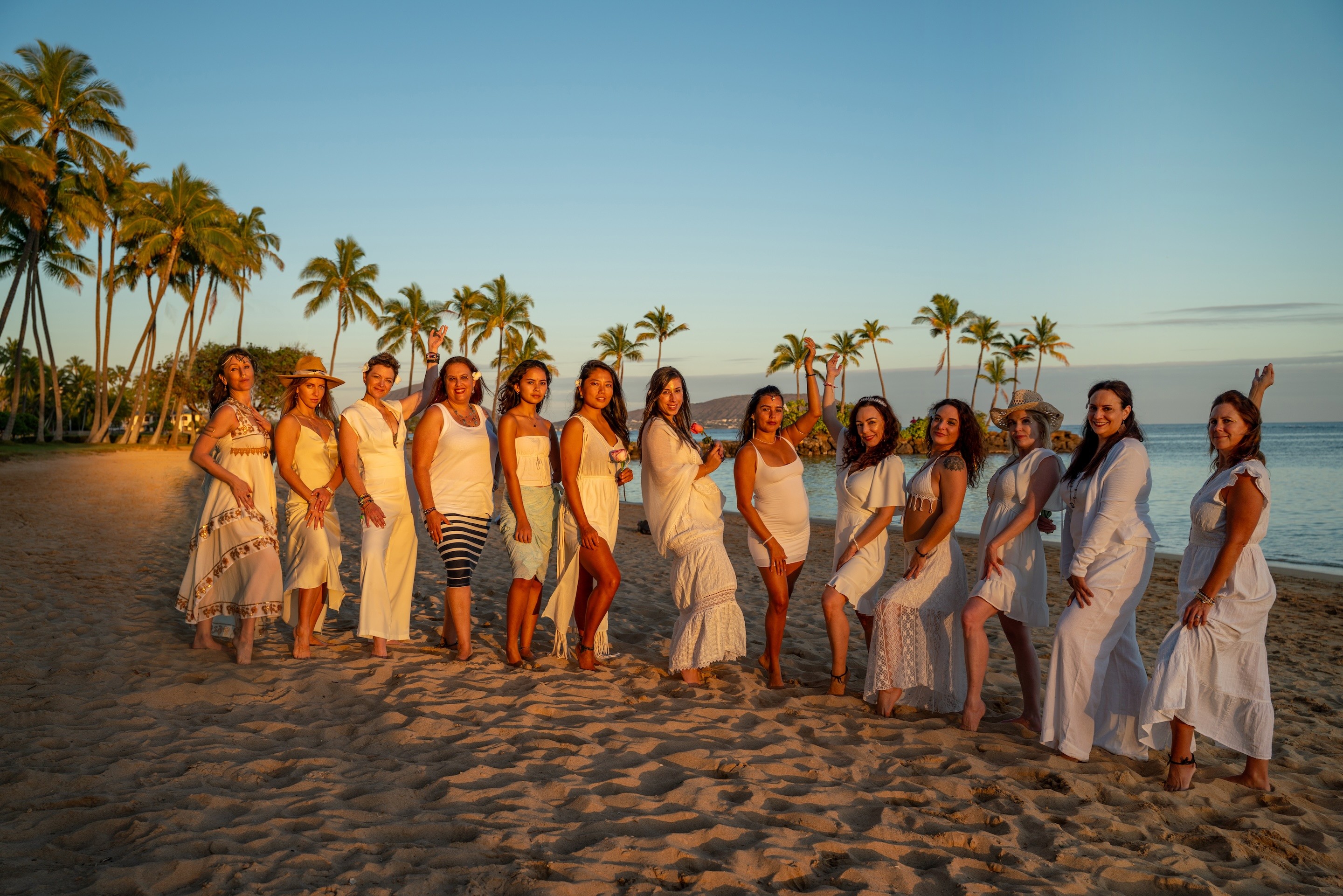 Group of women in white dresses standing on sandy beach with palm trees and blue sky in background.