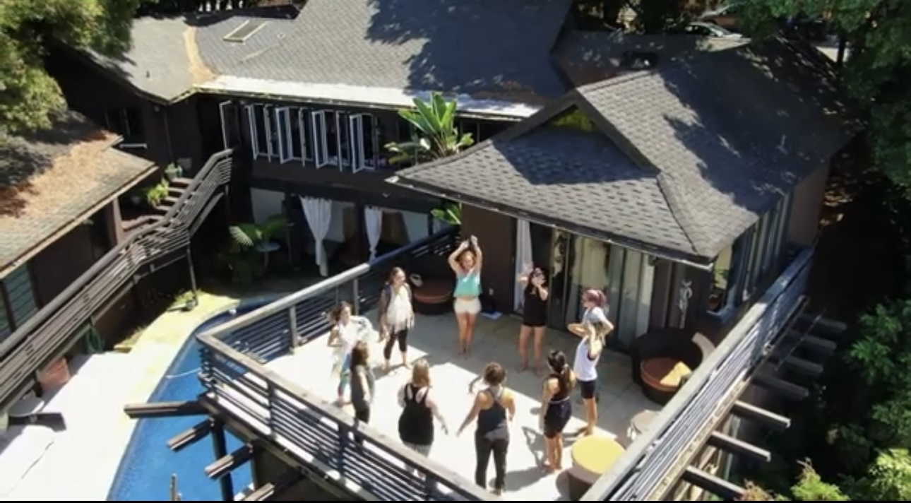 People gather on a rooftop deck with a pool, surrounded by railings and outdoor furniture, in a sunny setting.