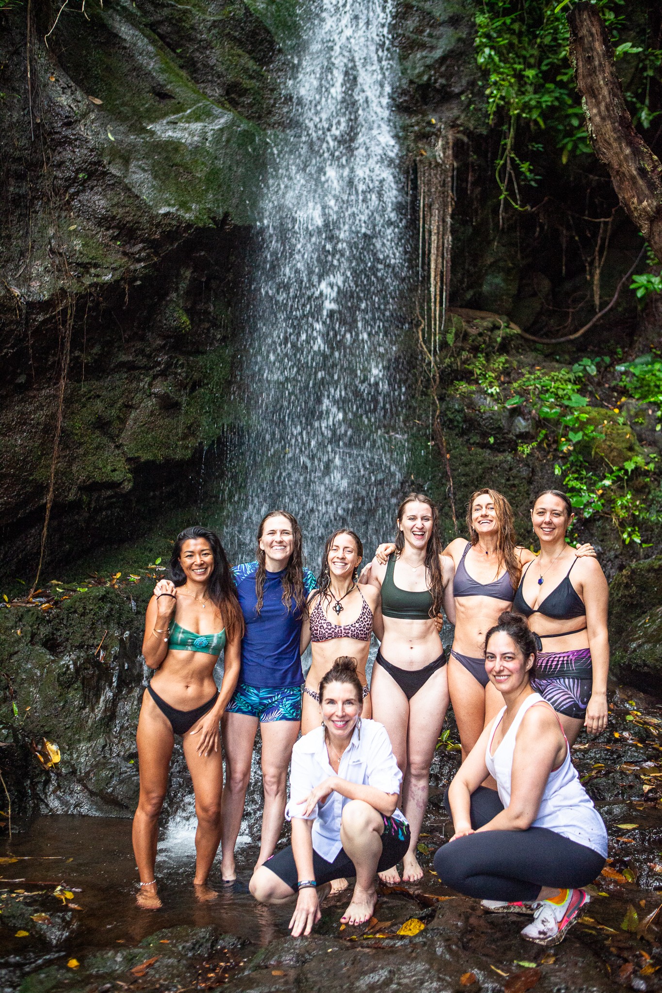 Group of eight women in swimsuits standing and kneeling in front of a waterfall in a forested area.