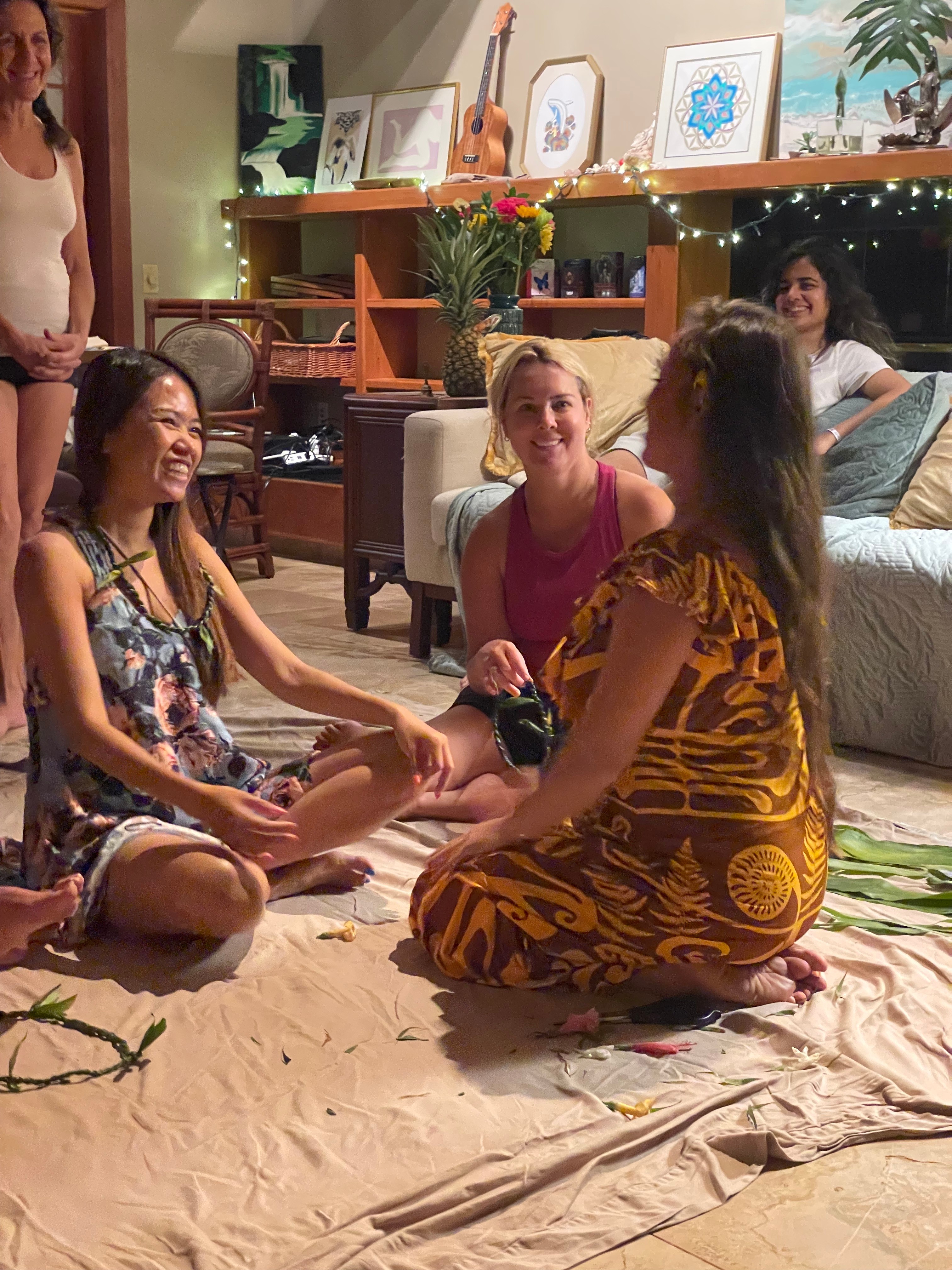 Four women sitting on a beige cloth on the floor, smiling and engaging in conversation in a cozy living room.