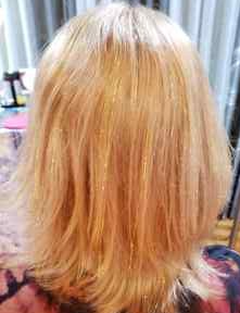 Back view of a person with shoulder-length, blonde hair, slightly wavy, in a room with curtains and furniture.