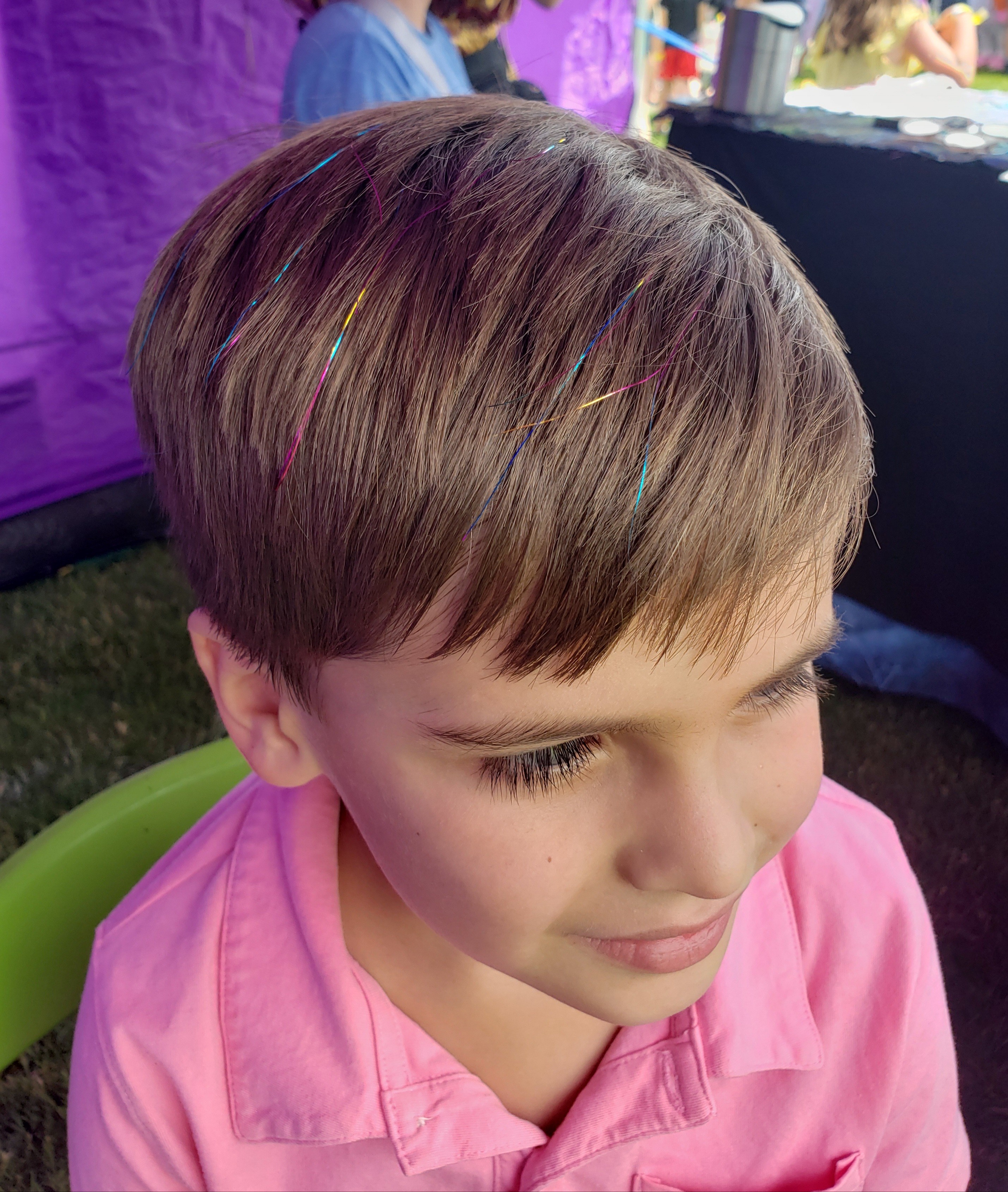 Young boy with brown hair wearing a pink shirt, looking downward, outdoors with purple and black background.