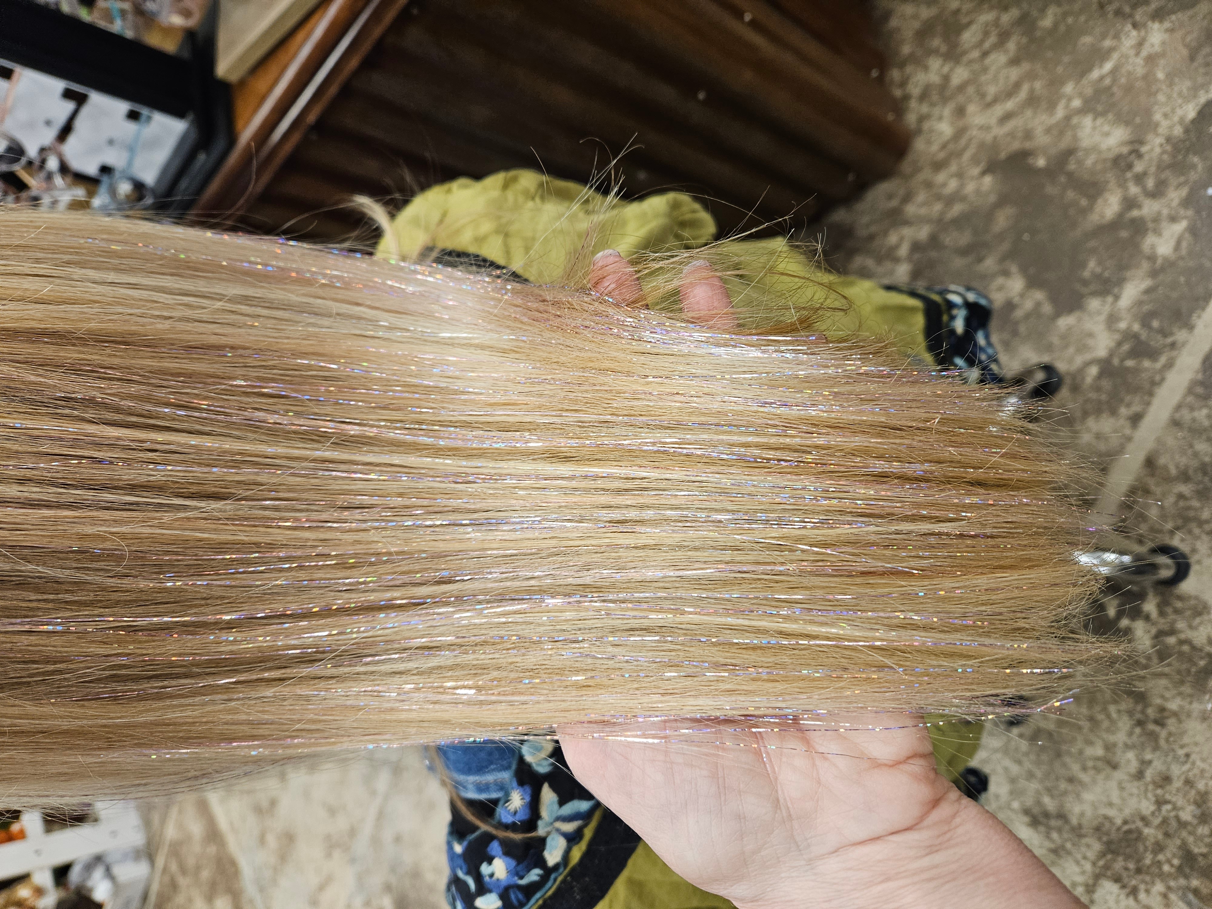 Person holding a bundle of long, straight, blonde hair with a hand visible, background shows a workshop or storage area.