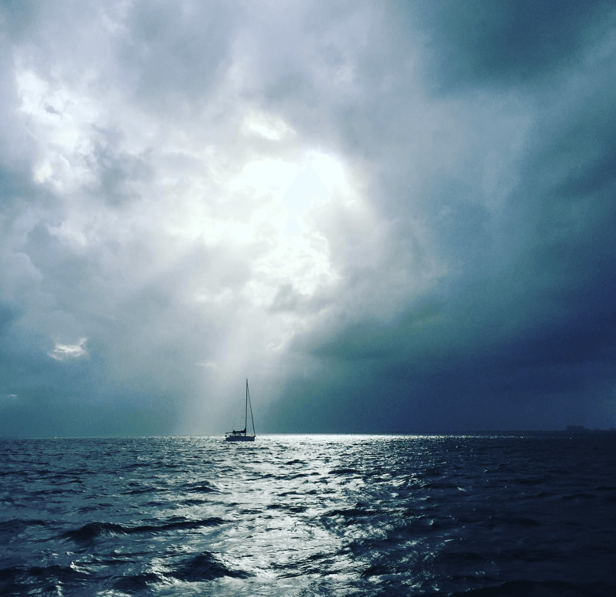 Sailboat on the ocean under a cloudy sky with sunlight breaking through, dark clouds overhead, and waves in the water.