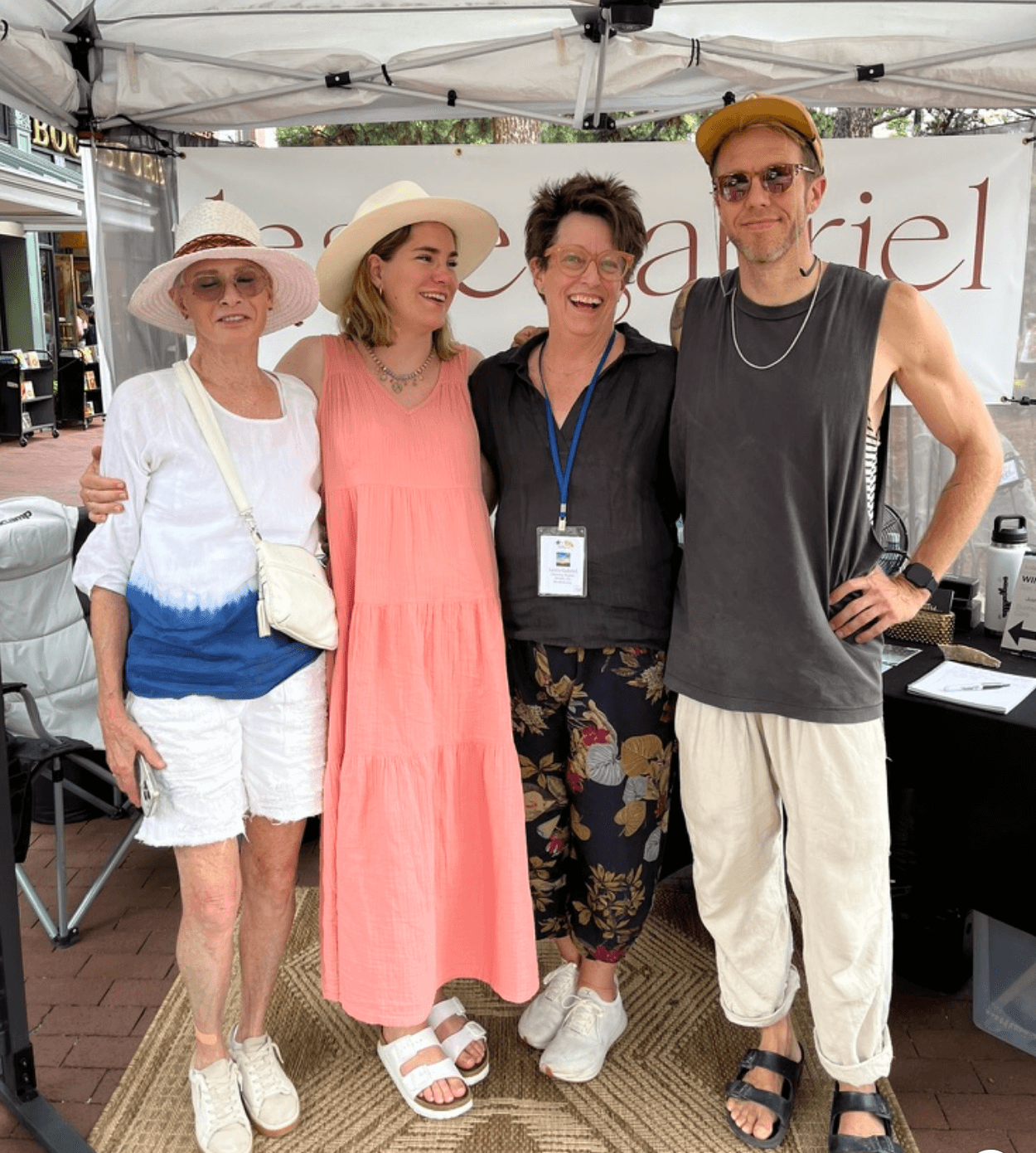 Four people standing together under a tent, smiling, with a banner in the background, dressed casually and wearing hats.