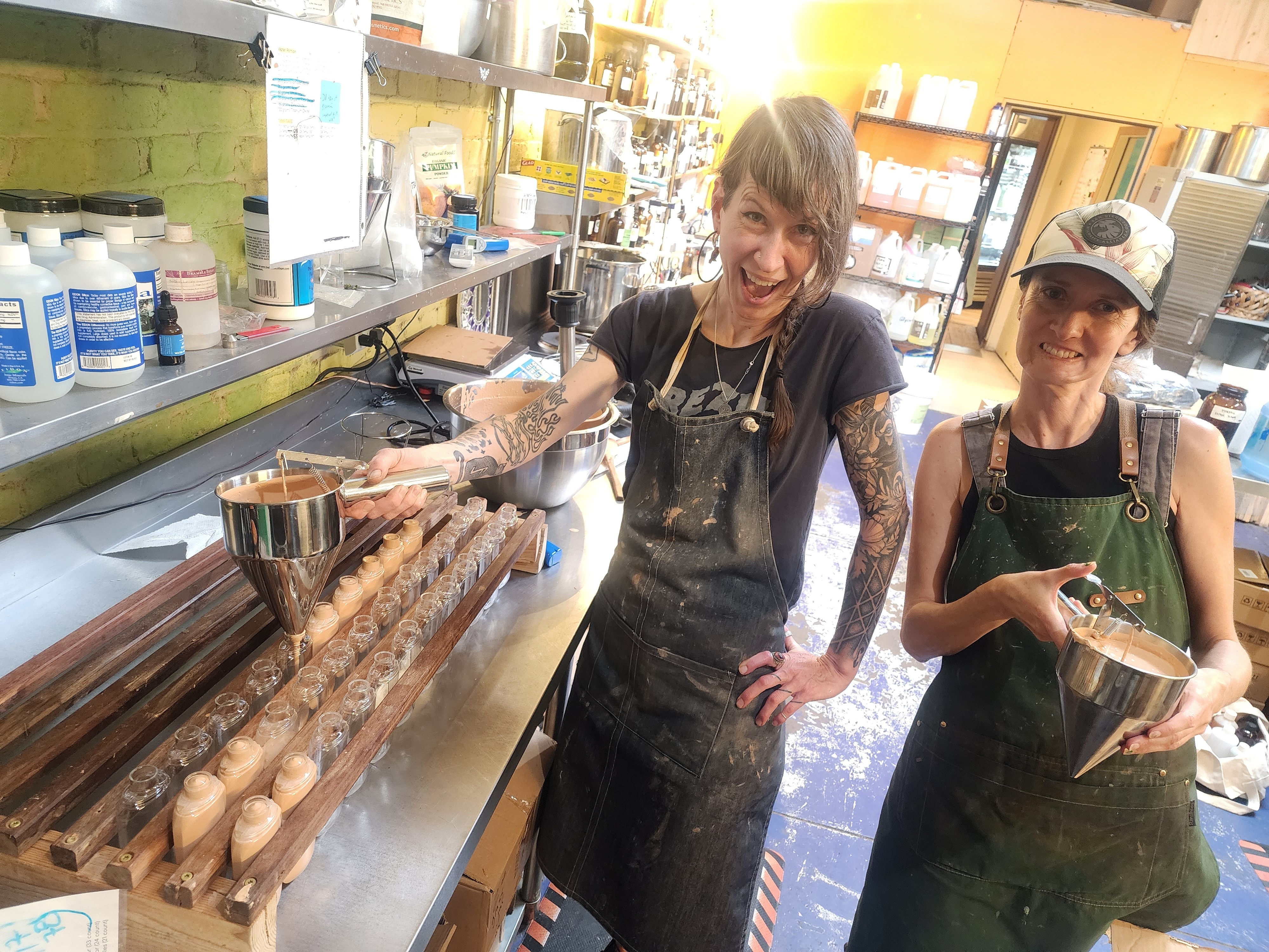 Two women in a kitchen, one smiling and holding a metal container, the other holding a mixing bowl, surrounded by kitchen supplies.