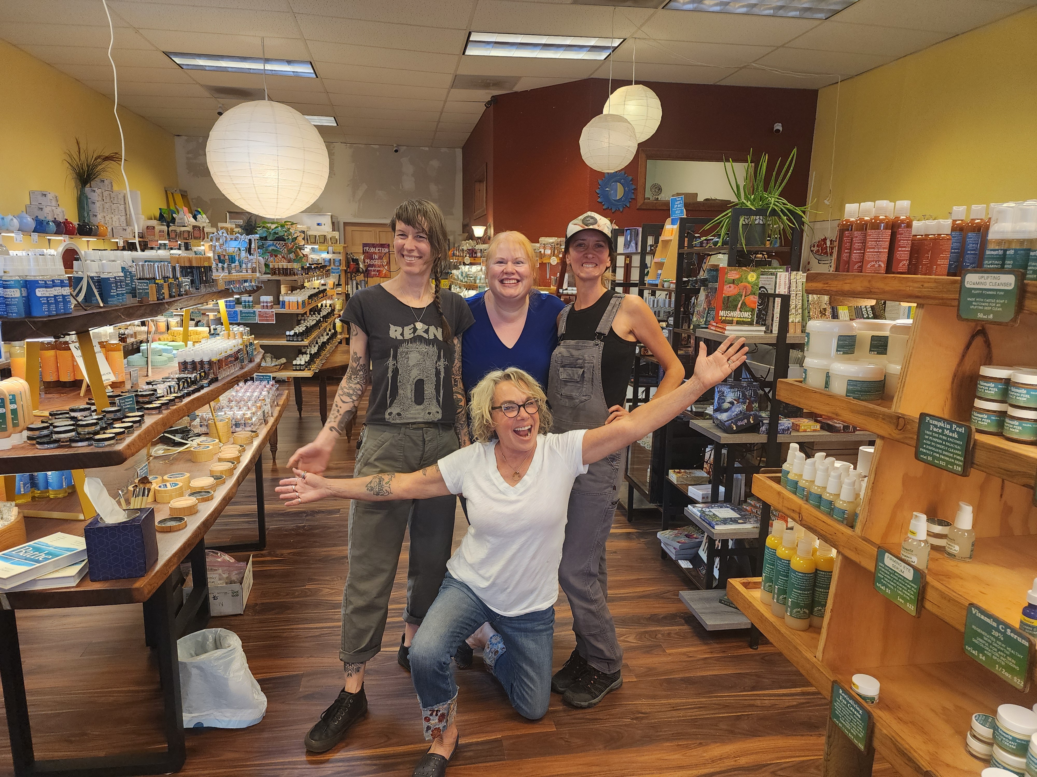 Four women in a store, smiling, with shelves of products behind them, one woman kneeling with arms outstretched.