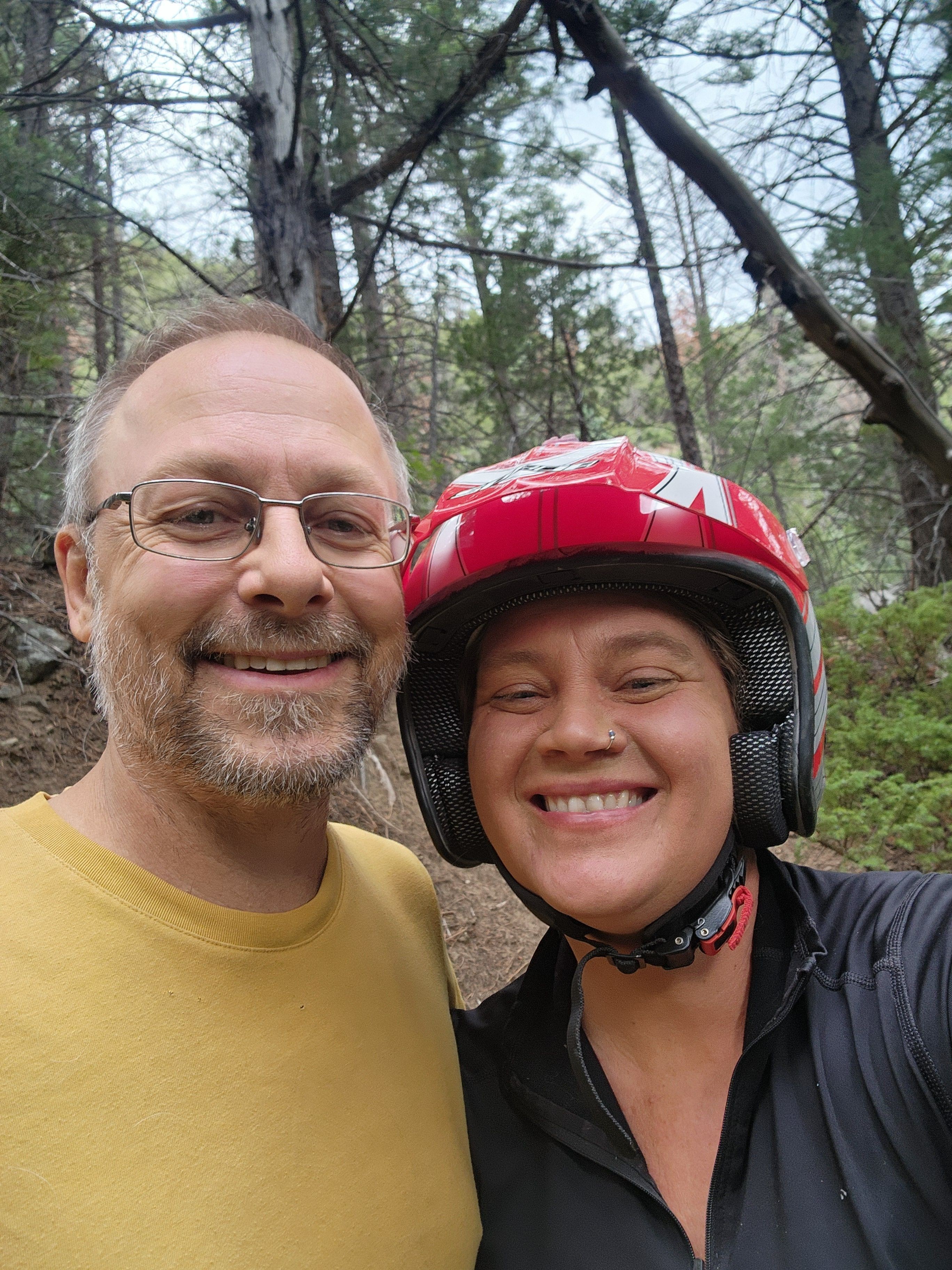 Two smiling people outdoors, one wearing glasses and a yellow shirt, the other with a helmet and black jacket, trees in background.