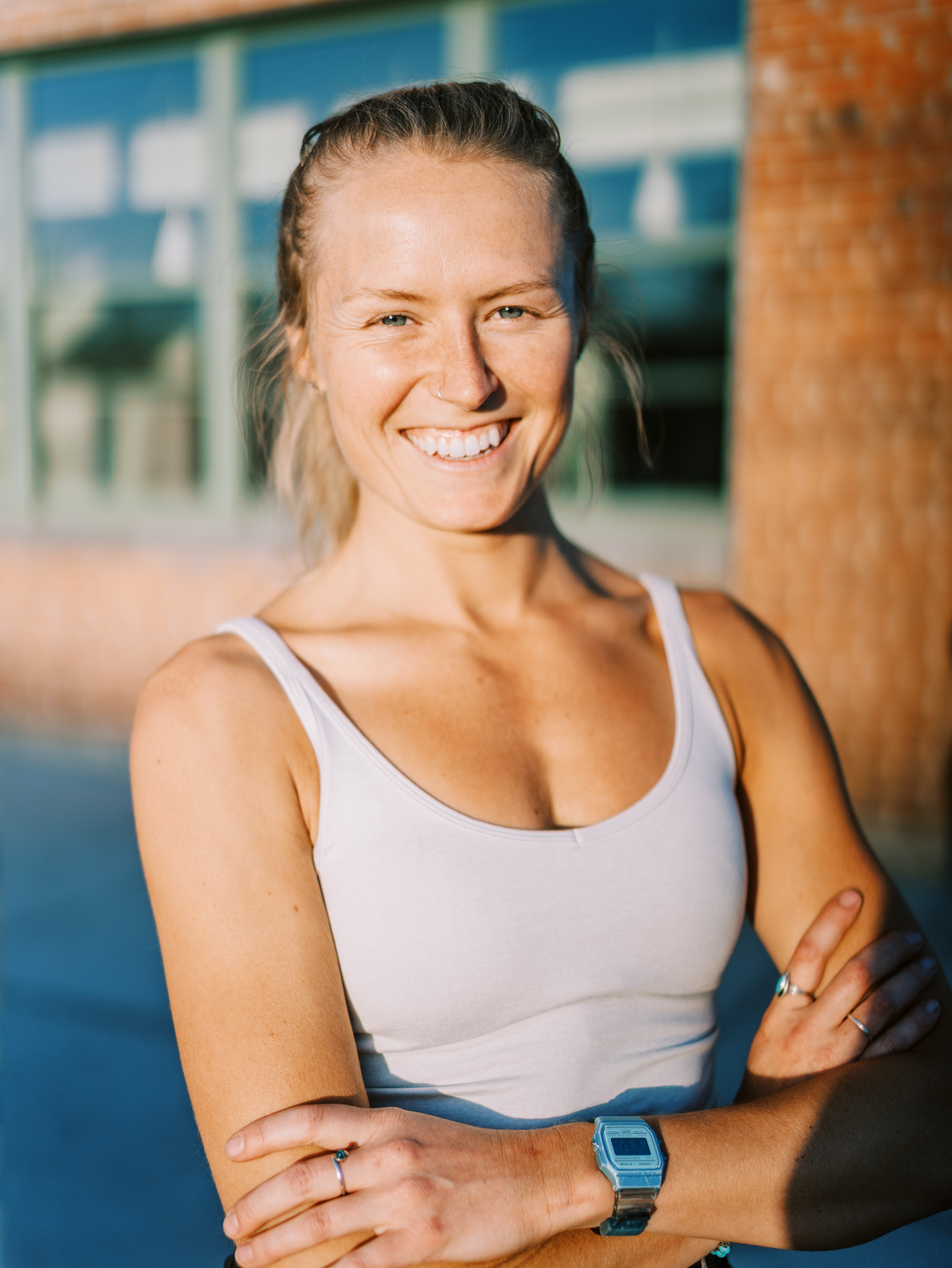 Smiling woman with blonde hair in a ponytail, wearing a white tank top and blue watch, standing outdoors.