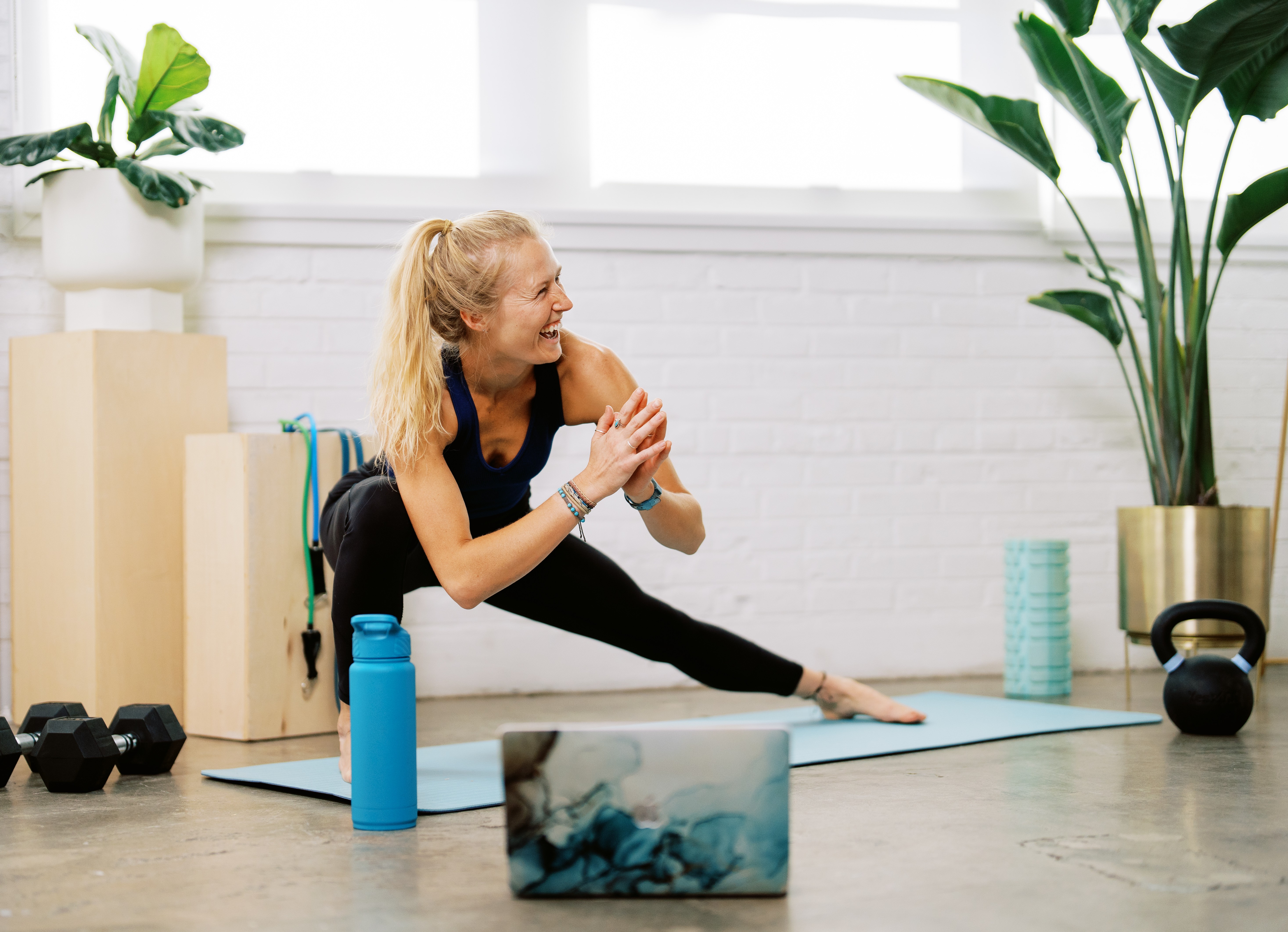 Woman stretching on yoga mat in a bright room with plants and workout equipment.