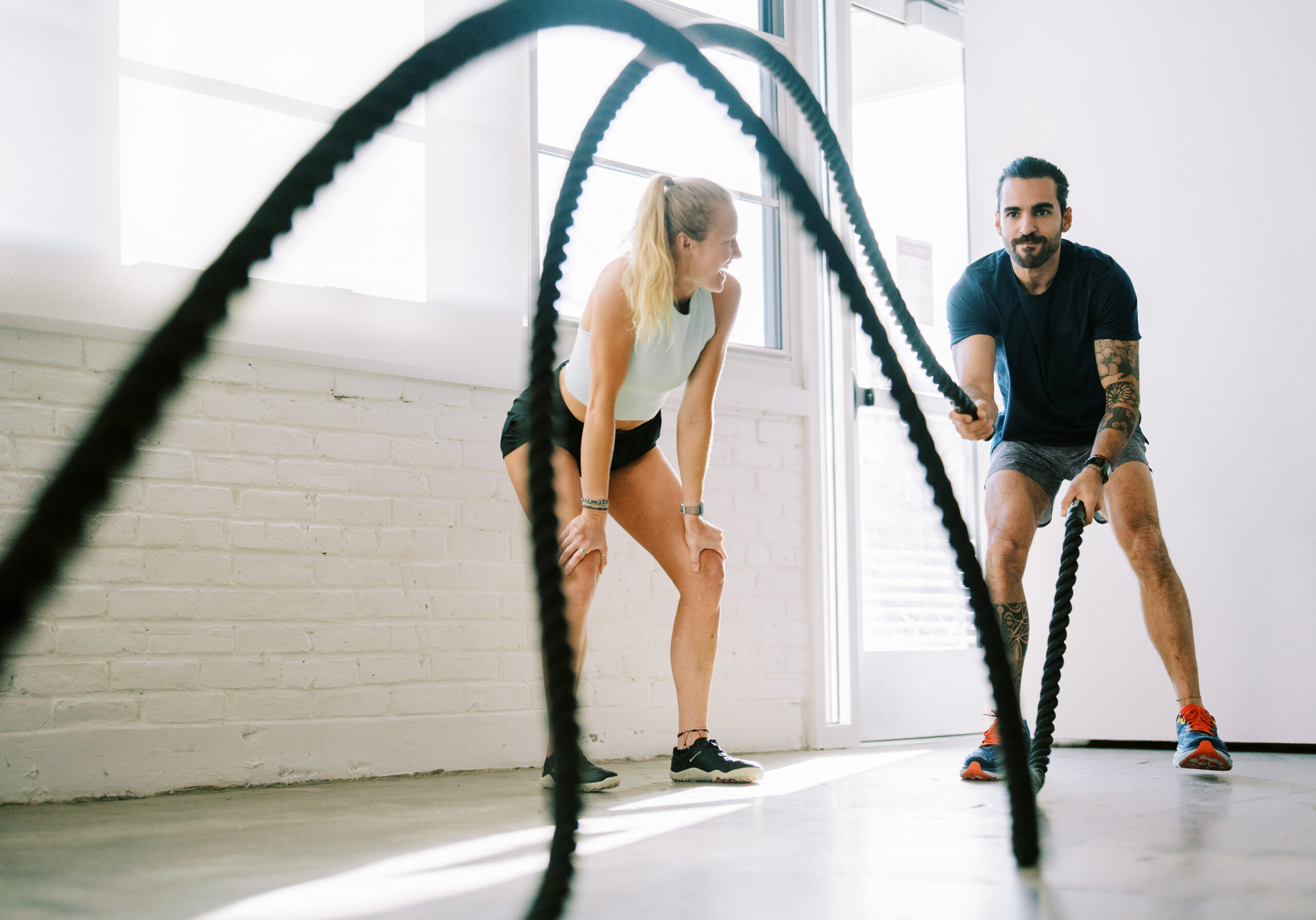 Two people exercising with battle ropes in a bright room, one woman and one man, both smiling and engaged.