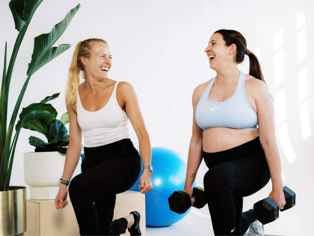 Two women laughing and exercising with dumbbells in a bright room with a large plant and a blue exercise ball.