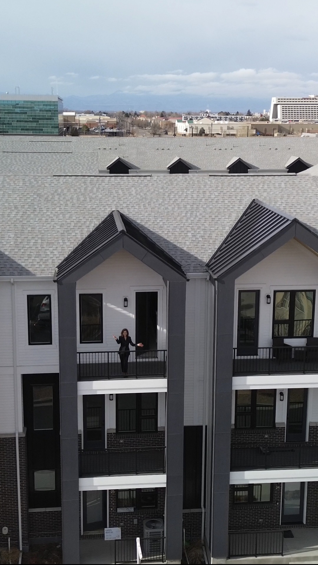 View of a modern building with balconies, a person standing on a balcony, and a cityscape in the background.
