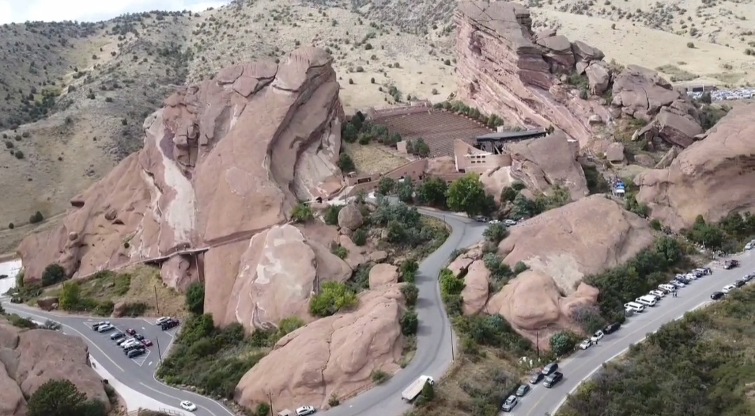 Rock formations with a winding road and parked cars at the base, surrounded by greenery and hills.