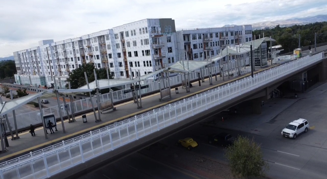 Elevated train station with platform, stairs, and people, above a street with cars and trees, modern apartment buildings in background.