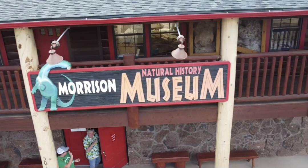 Sign for Morrison Natural History Museum with cartoon character, mounted on a building with a red railing and stone wall.