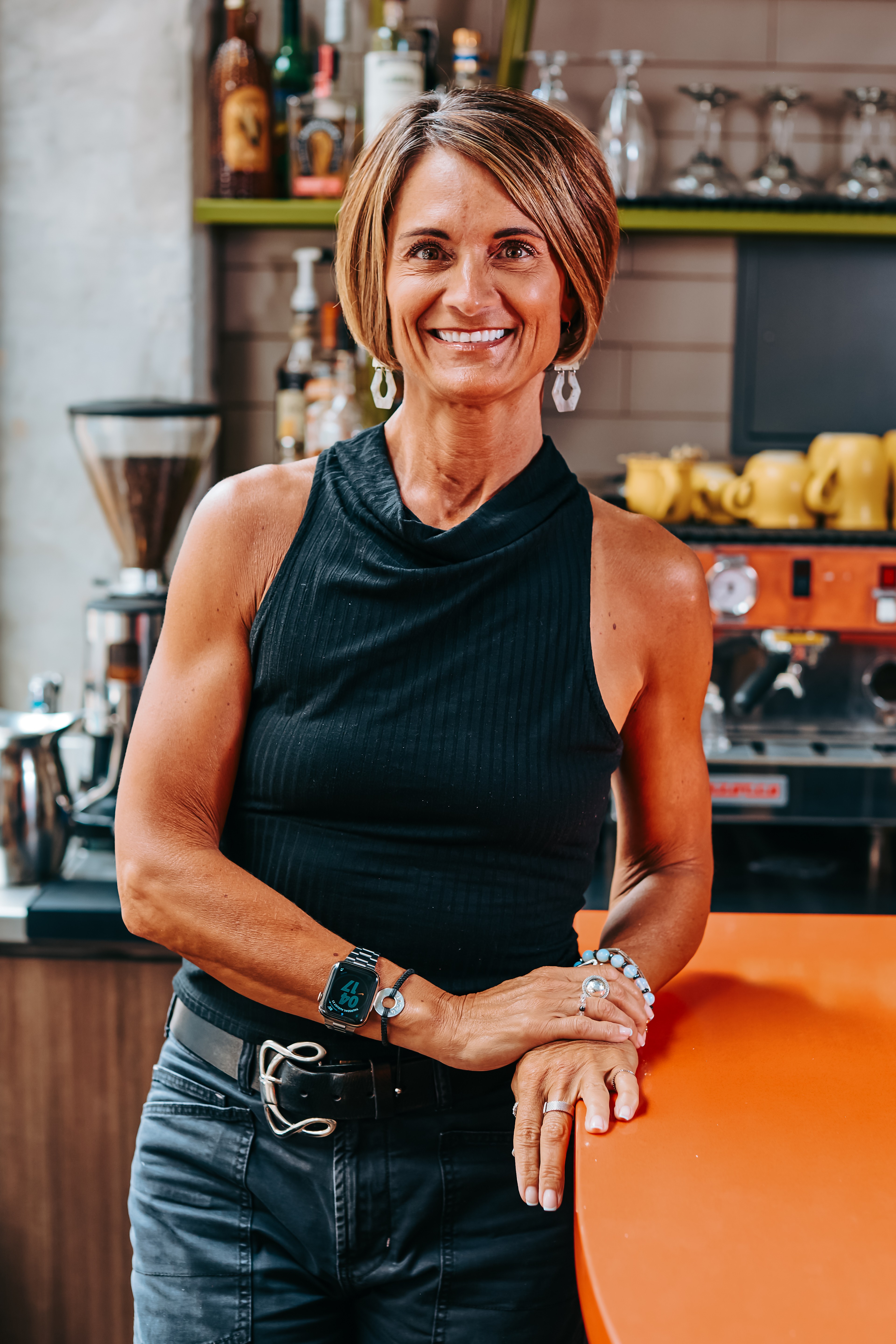 Smiling woman with short hair in a sleeveless black top standing in a bar or cafe setting.