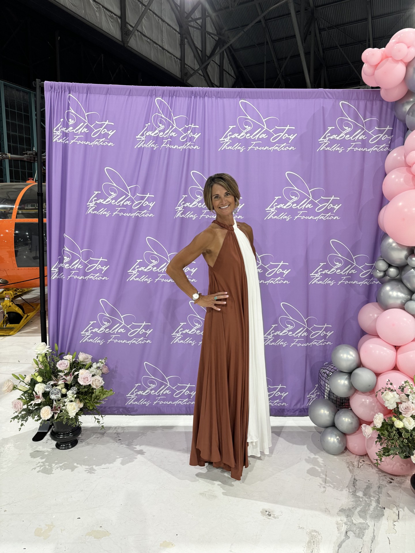 Woman in a brown and white dress standing in front of a purple backdrop with floral decorations.