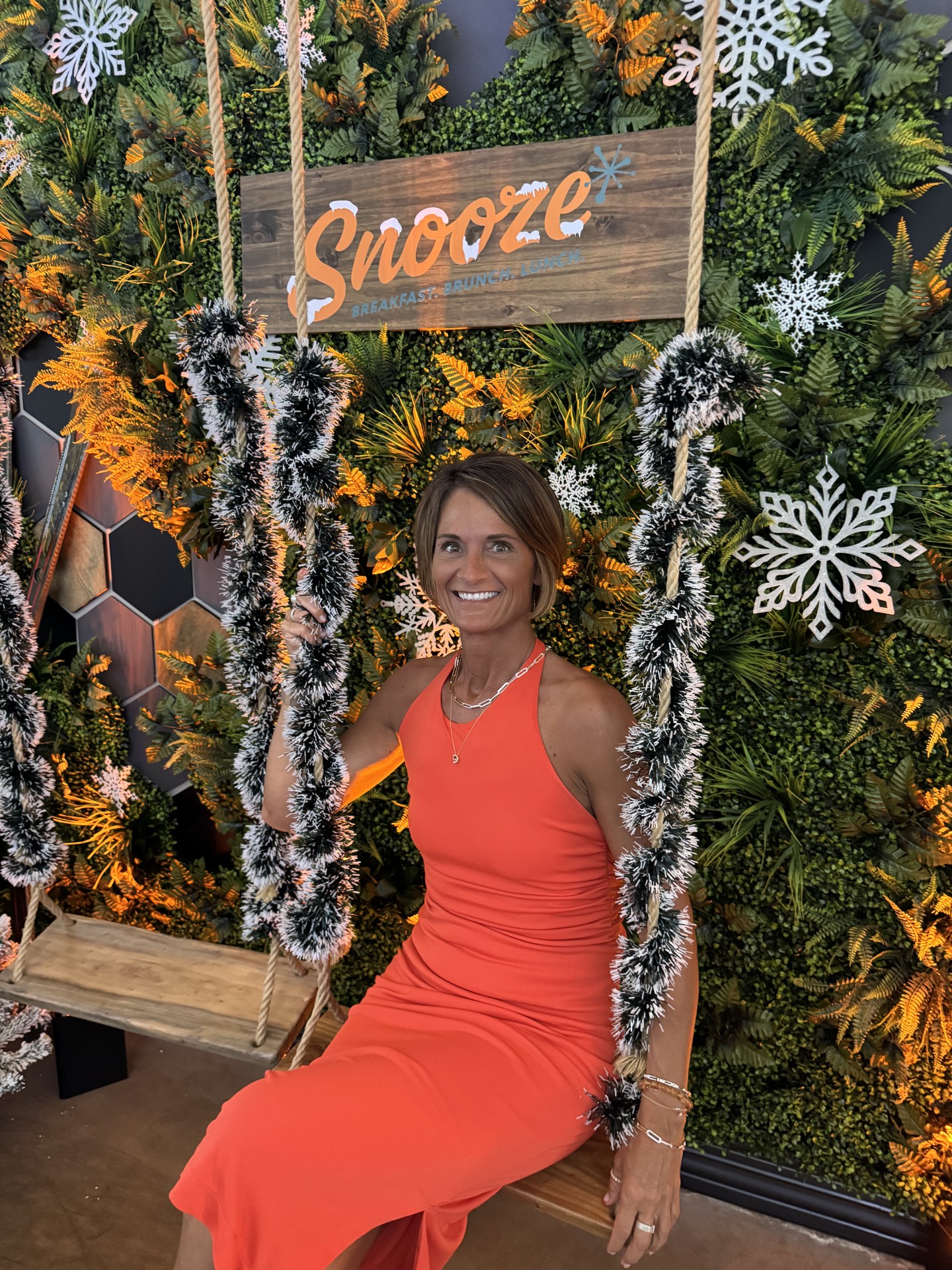Woman in orange dress sitting on a decorated swing with a festive background, smiling, surrounded by holiday decorations.