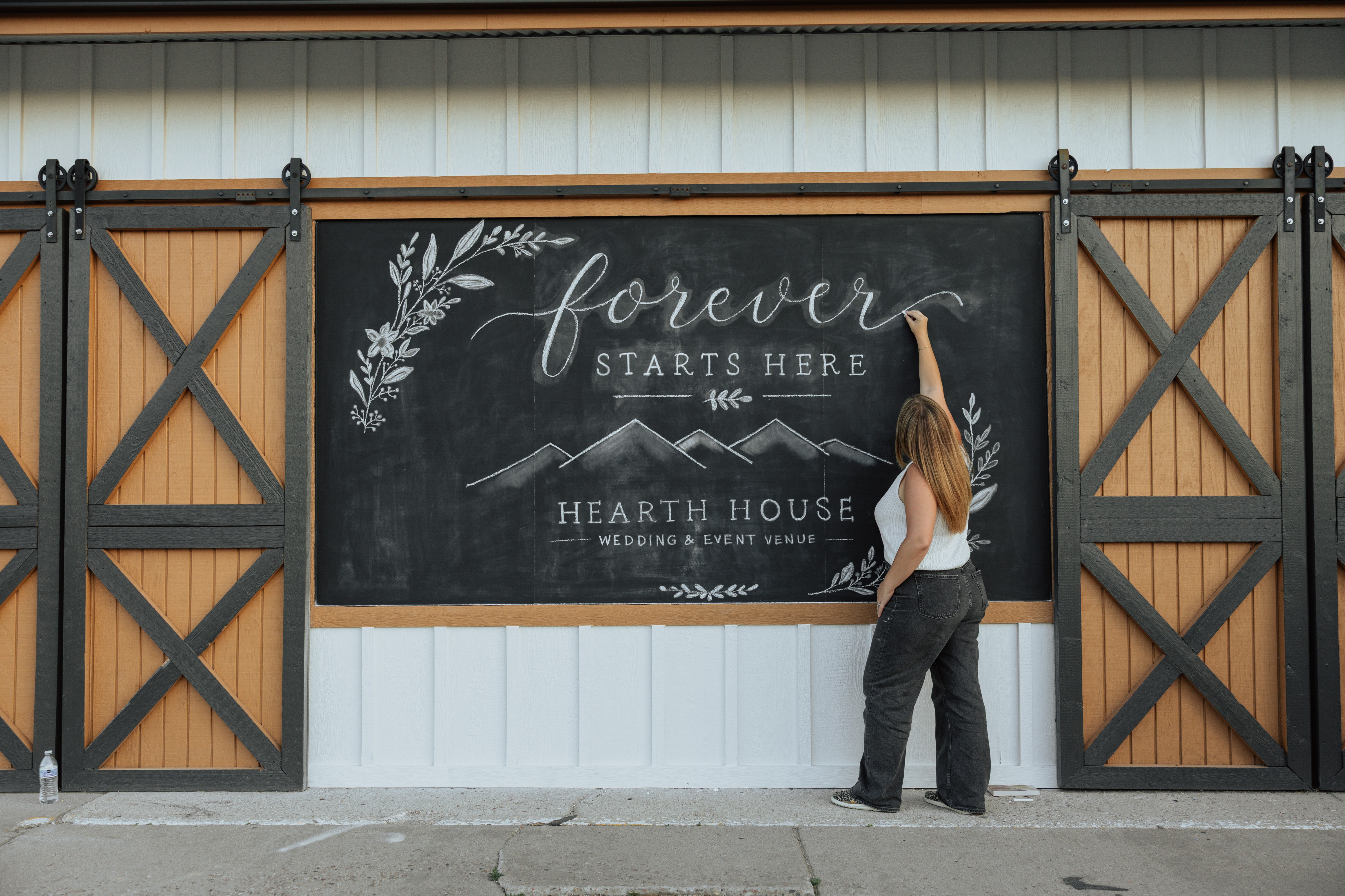 Person writing on a large blackboard with the words 'forever starts here' and 'Hearth House' in a barn-like setting.