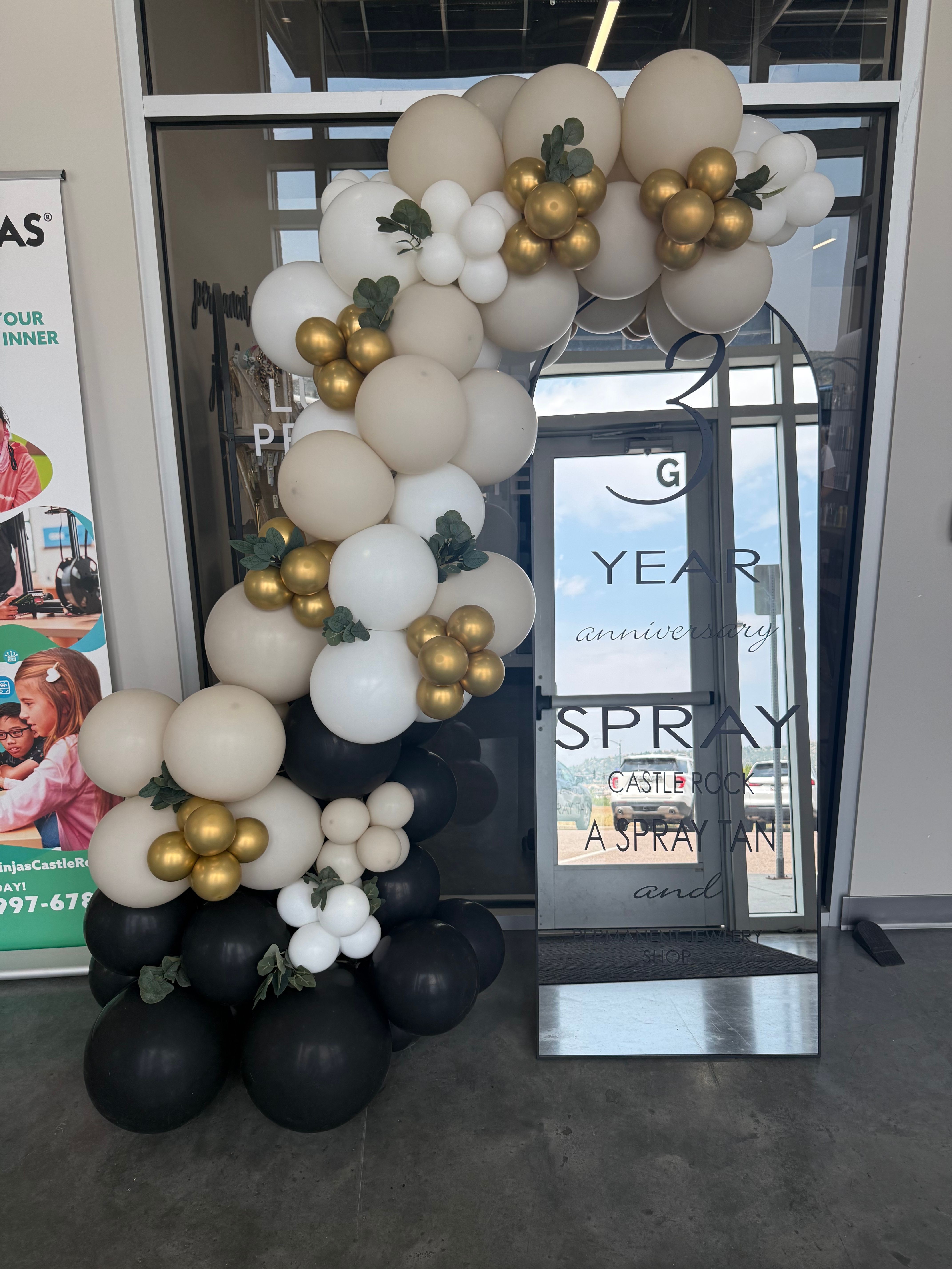 Decorative balloon arrangement with white, black, and gold balloons in front of a glass door, with store signs visible behind.
