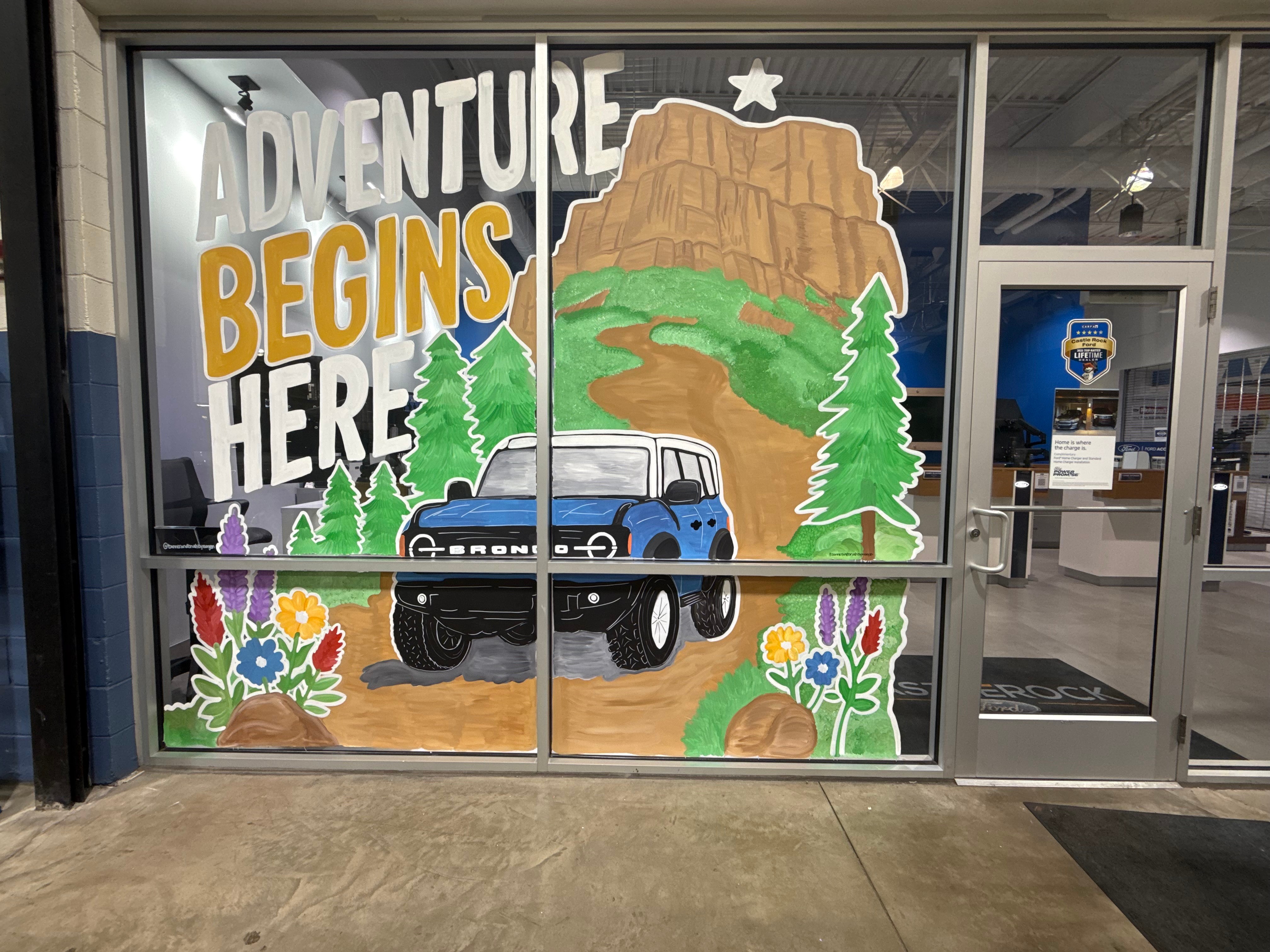 Decorative window with a mountain, trees, a vehicle, and colorful flowers, with the text 'Adventure Begins Here'.