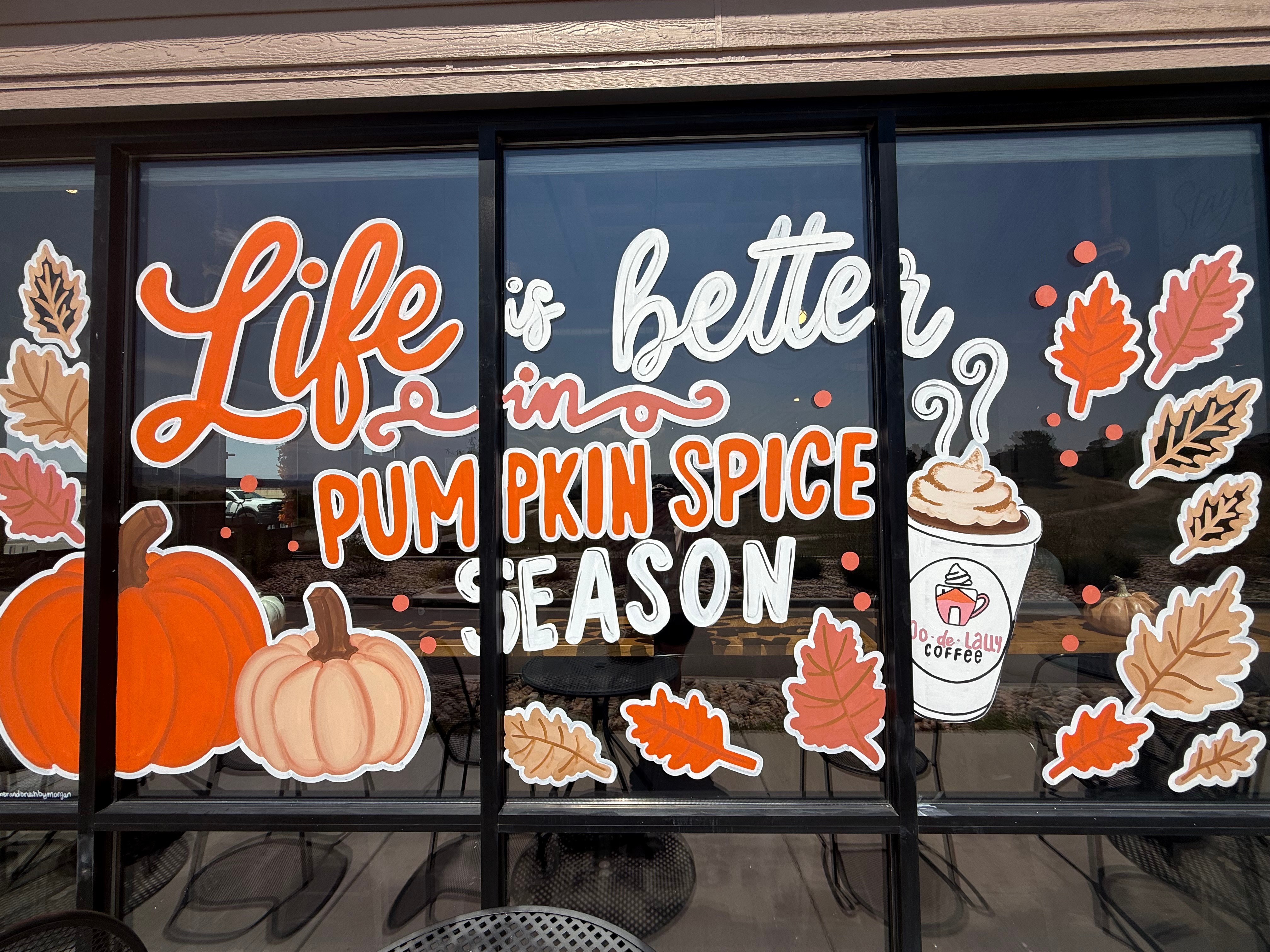 Window display with fall-themed decorations, pumpkins, leaves, and a coffee cup, promoting pumpkin spice season.