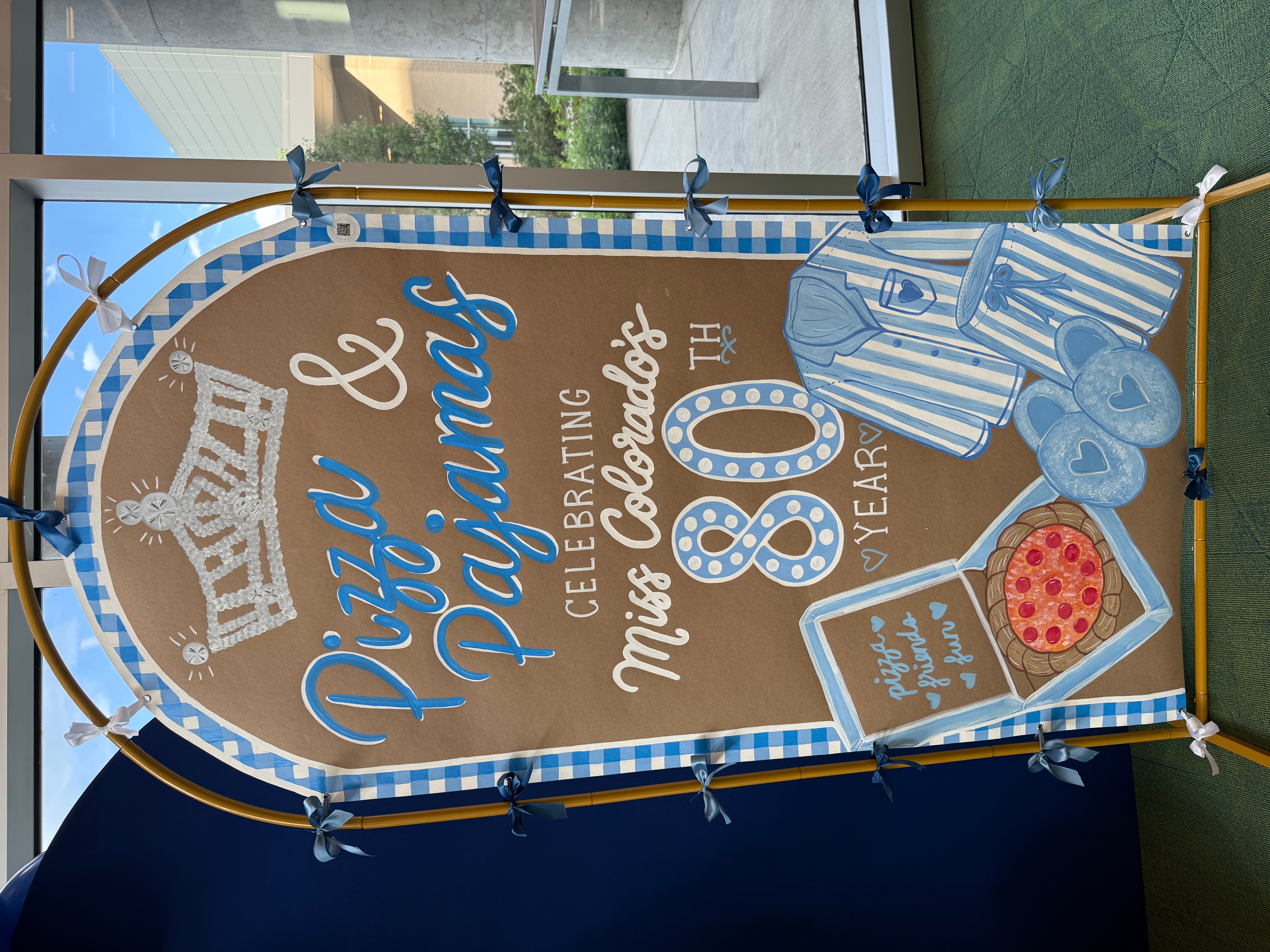 Celebration sign with a pizza, striped shirt, and text celebrating an 80th birthday, displayed indoors near windows.