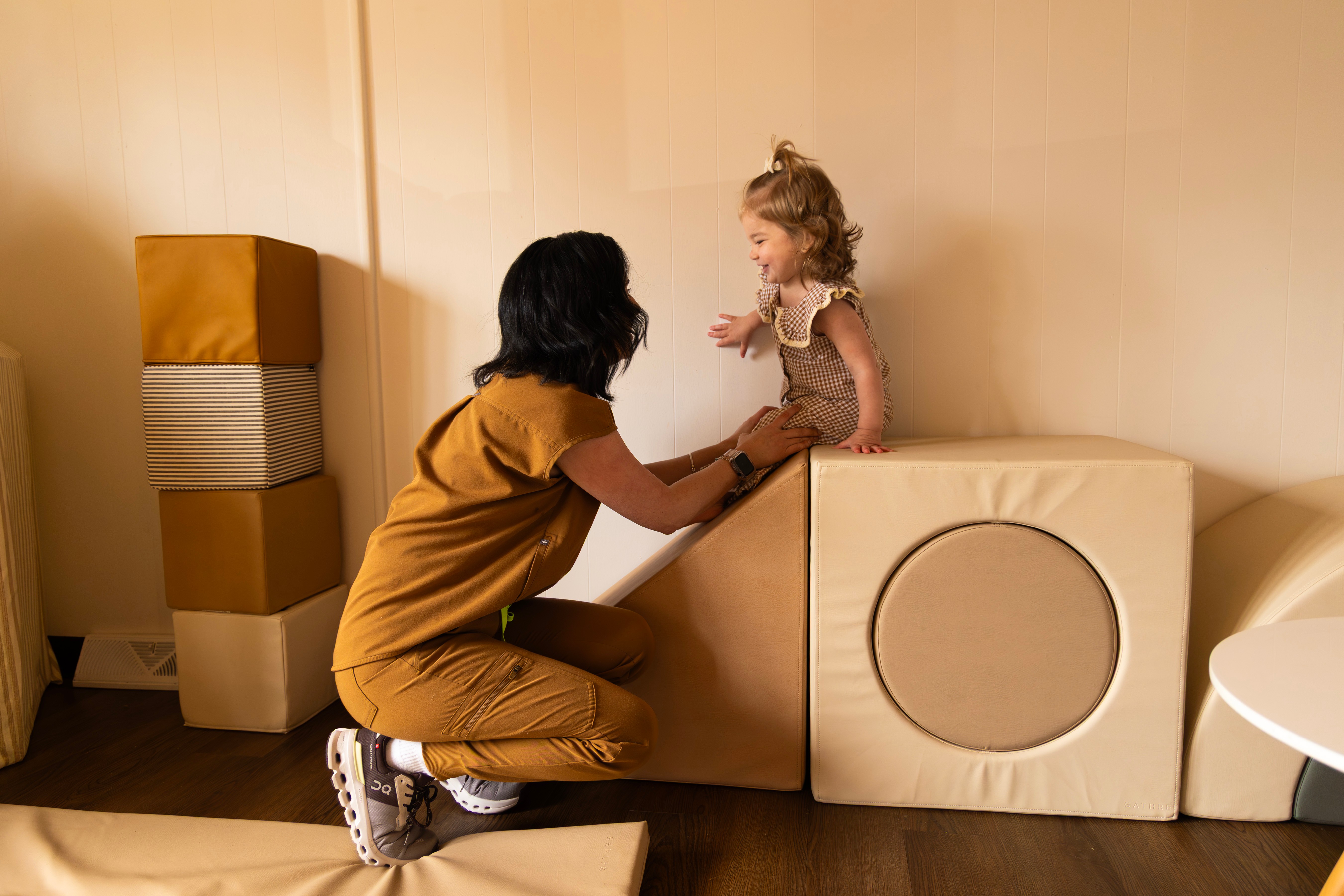 Adult woman kneeling and holding hands with a young girl standing on a block in a room with beige walls and furniture.