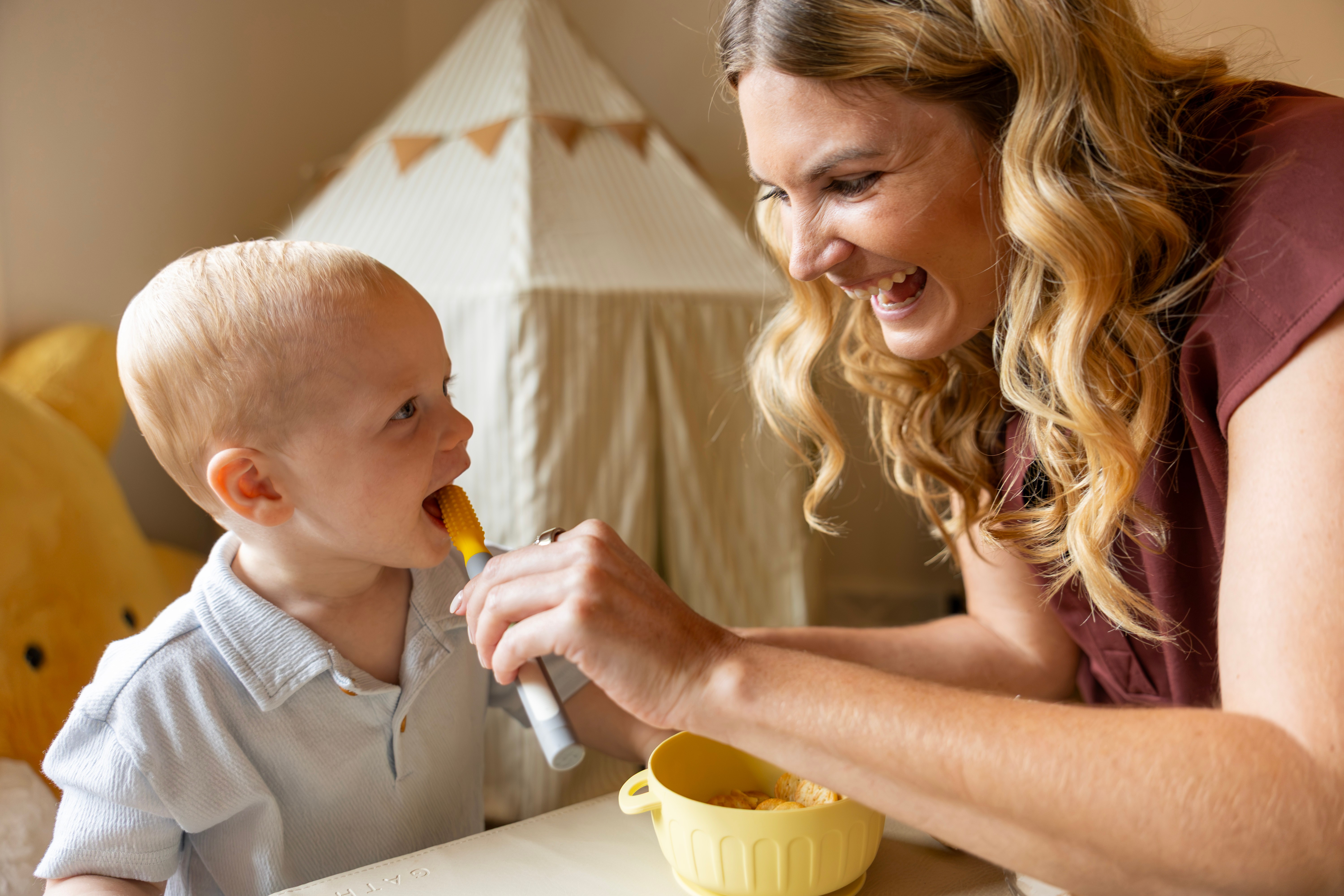 Woman feeding a young child with a spoon, both smiling, in a cozy room with a tent in the background.